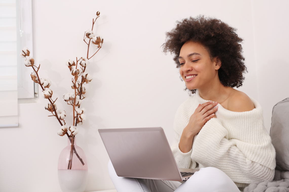 Woman with curly black hair sits on a couch in her home, her laptop is on her lap while she is in a telehealth appointment