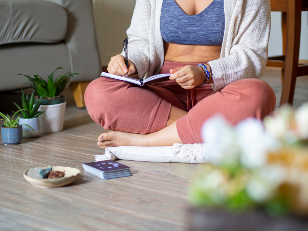 A woman sits on a yoga mat in her home with a journal on her lap