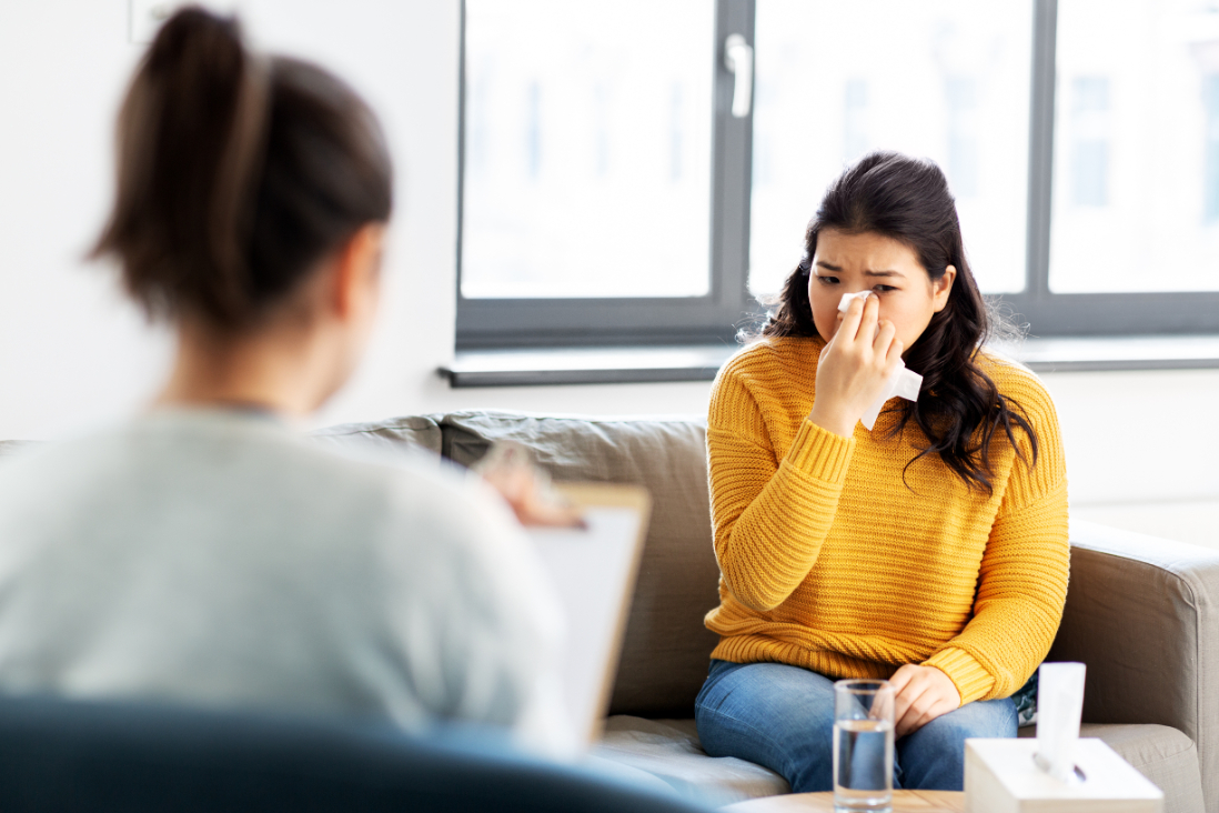 A woman in a yellow shirt sits in front of a therapist while holding a tissue to her eye to stop a tear