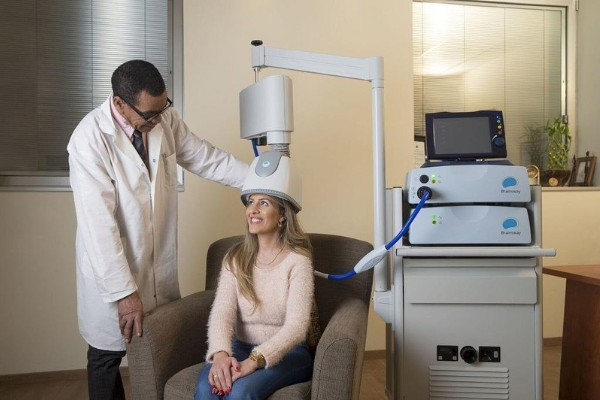 Woman receiving TMS treatment with helmet and machine pictured. Doctor standing next to patient. 