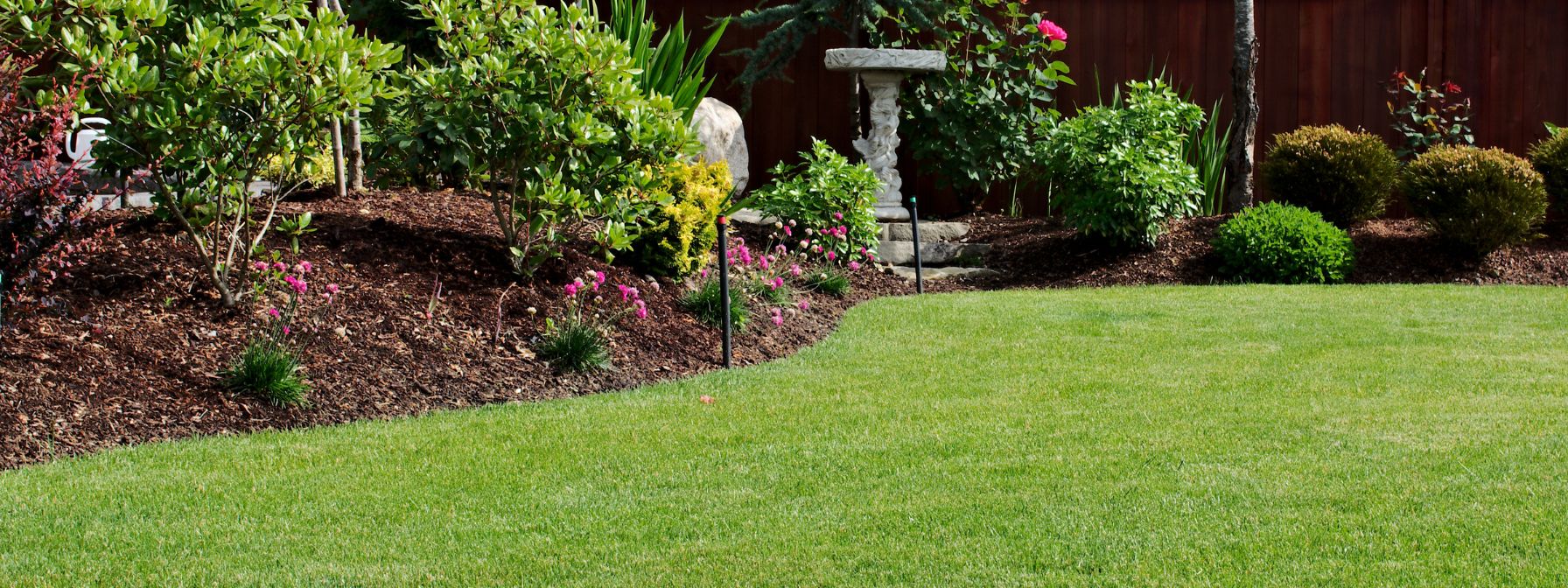 Well-maintained backyard featuring a lush green lawn bordered by mulched garden beds with shrubs, flowers, and ornamental elements—illustrating the results of healthy soil and effective landscaping practices. Well-maintained backyard featuring a lush green lawn bordered by mulched garden beds with shrubs, flowers, and ornamental elements—illustrating the results of healthy soil and effective landscaping practices.
