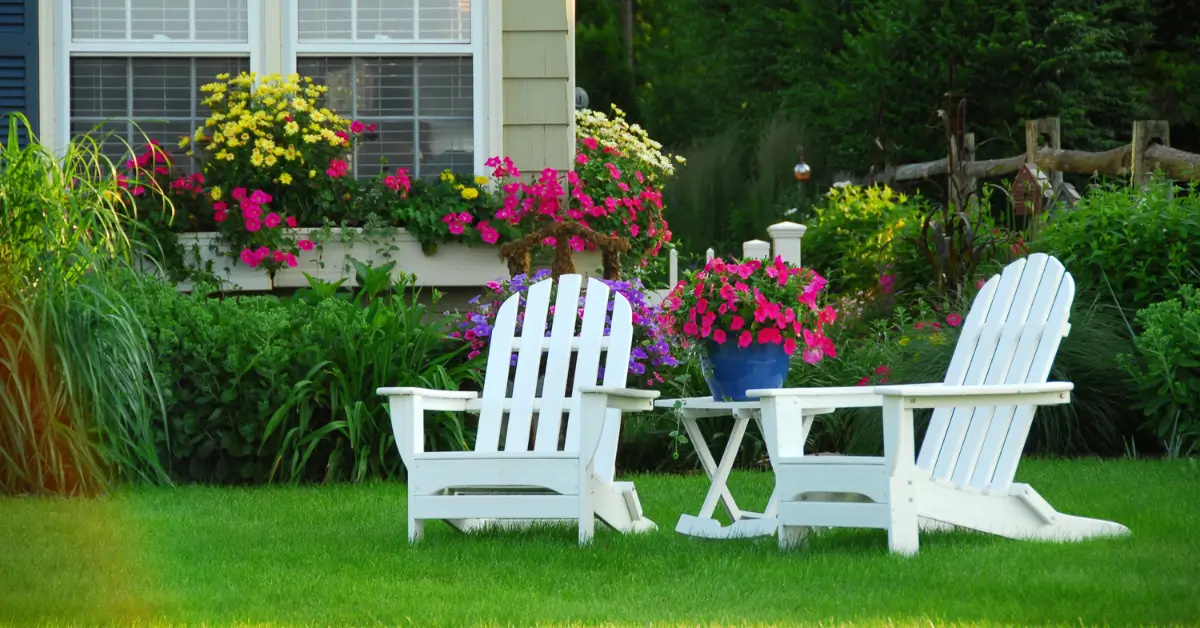 Two white Adirondack chairs with a small table between them sit on a well-manicured lawn, surrounded by vibrant flowers in pots and window boxes in front of a house. Two white Adirondack chairs with a small table between them sit on a well-manicured lawn, surrounded by vibrant flowers in pots and window boxes in front of a house.