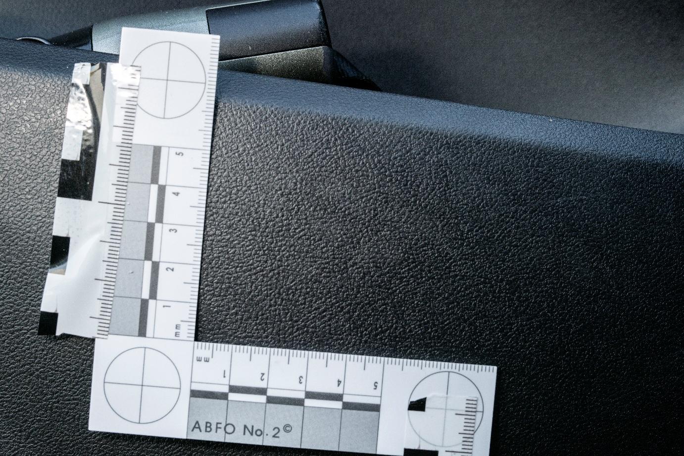 Close-up image of a textured black plastic surface inside a vehicle, showing two ABFO No. 2 forensic measurement scales arranged in an L-shape. The scales are secured with reflective black-and-white tape and positioned to measure evidence on the door trim panel. The photo appears to document a specific area for forensic comparison or analysis. Close-up image of a textured black plastic surface inside a vehicle, showing two ABFO No. 2 forensic measurement scales arranged in an L-shape. The scales are secured with reflective black-and-white tape and positioned to measure evidence on the door trim panel. The photo appears to document a specific area for forensic comparison or analysis.