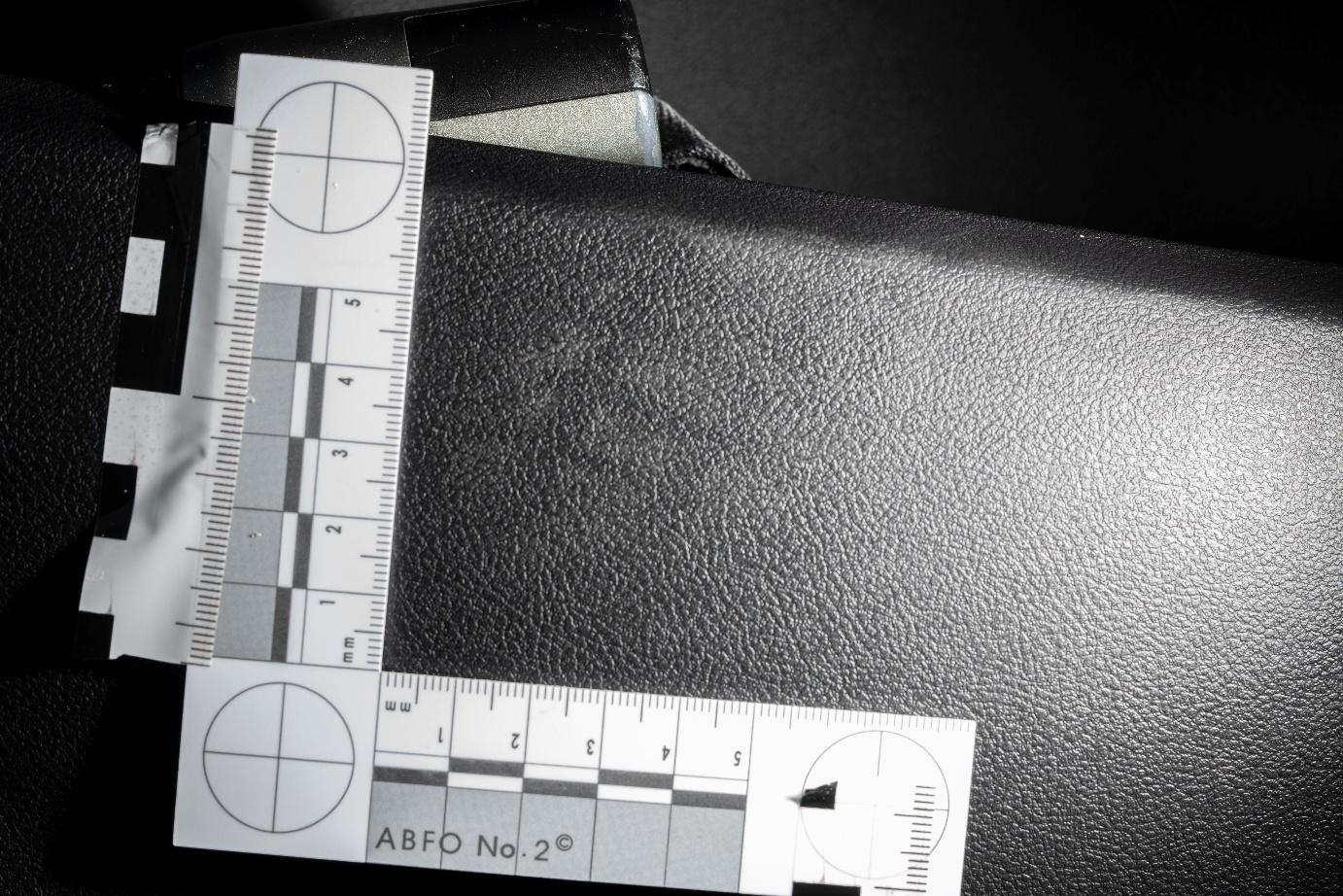 Close-up forensic image of a textured black plastic car interior surface with a partial footwear impression faintly visible in the center. Two ABFO No. 2 forensic measurement scales are arranged in an L-shape and secured with reflective evidence tape, aiding in the documentation of the mark's location and size. Bright directional lighting enhances the visibility of the impression. Close-up forensic image of a textured black plastic car interior surface with a partial footwear impression faintly visible in the center. Two ABFO No. 2 forensic measurement scales are arranged in an L-shape and secured with reflective evidence tape, aiding in the documentation of the mark's location and size. Bright directional lighting enhances the visibility of the impression.