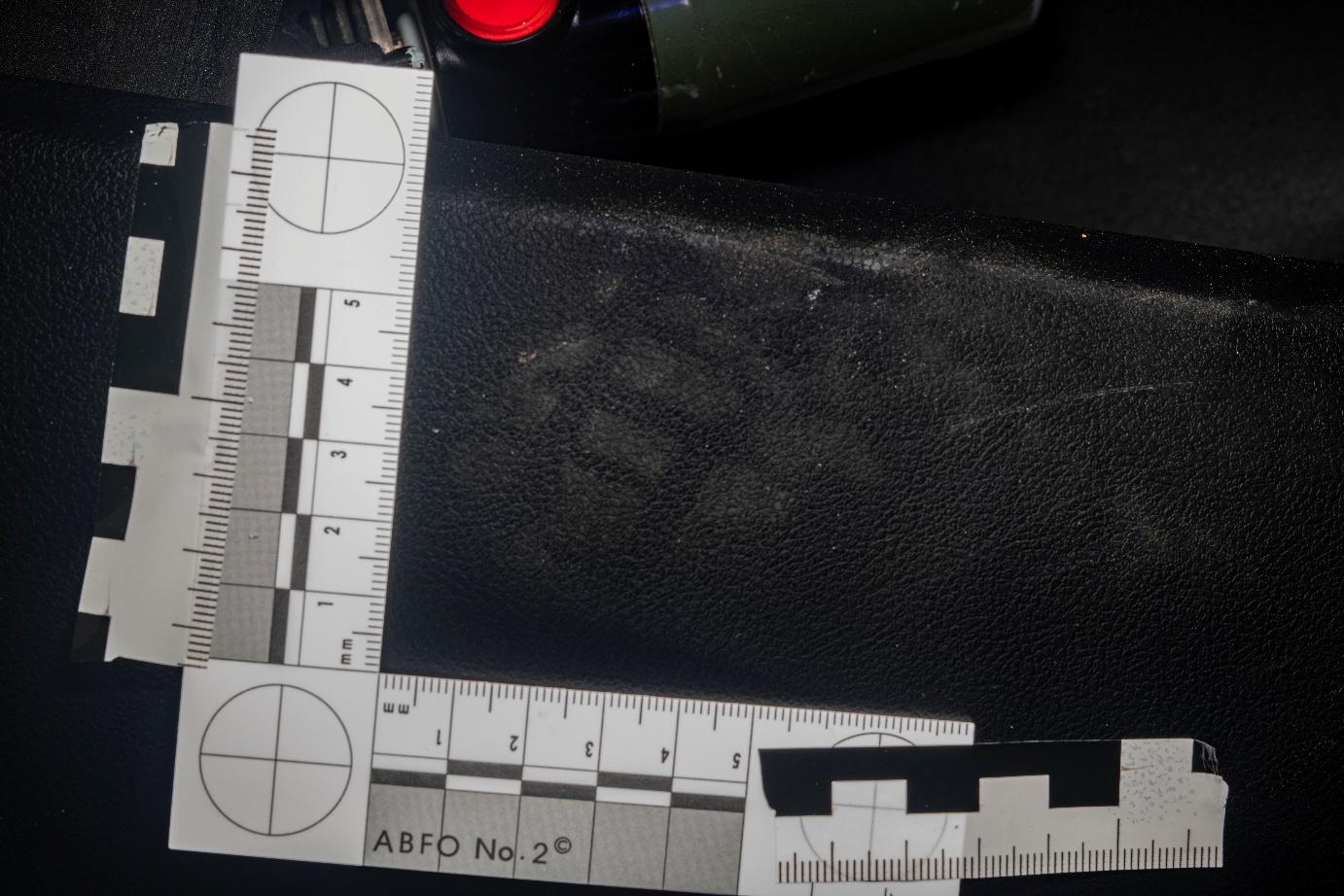 Forensic close-up image of a partial shoeprint mark on a black textured vehicle interior surface, documented using ABFO No. 2 measurement scales in an L-configuration. The mark is faint but visible in the center, enhanced by oblique lighting. Forensic close-up image of a partial shoeprint mark on a black textured vehicle interior surface, documented using ABFO No. 2 measurement scales in an L-configuration. The mark is faint but visible in the center, enhanced by oblique lighting.