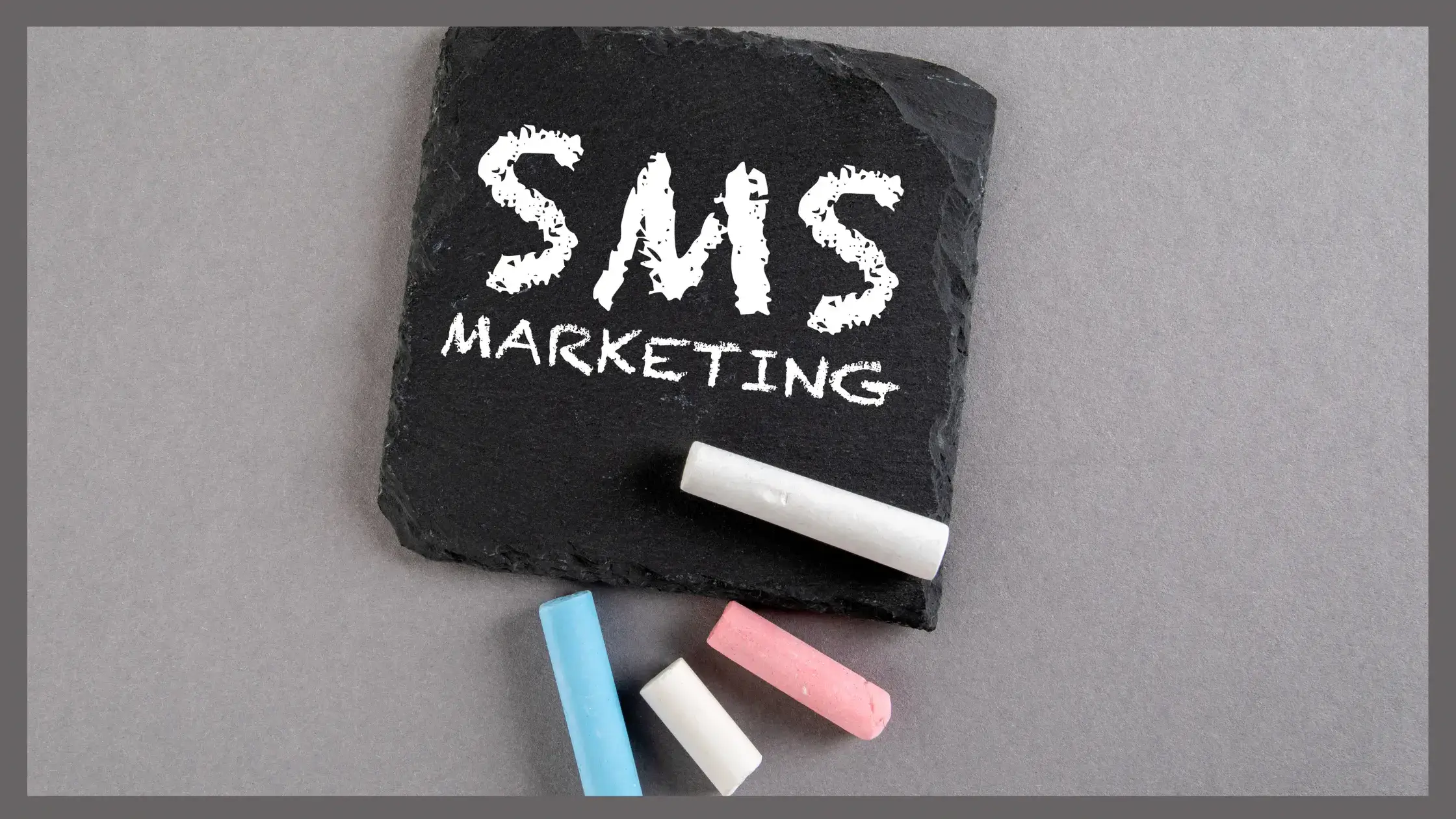 Image of a square black stone with white chalk written SMS Marketing, and 4 other pieces of chalk in blue pink and white colors