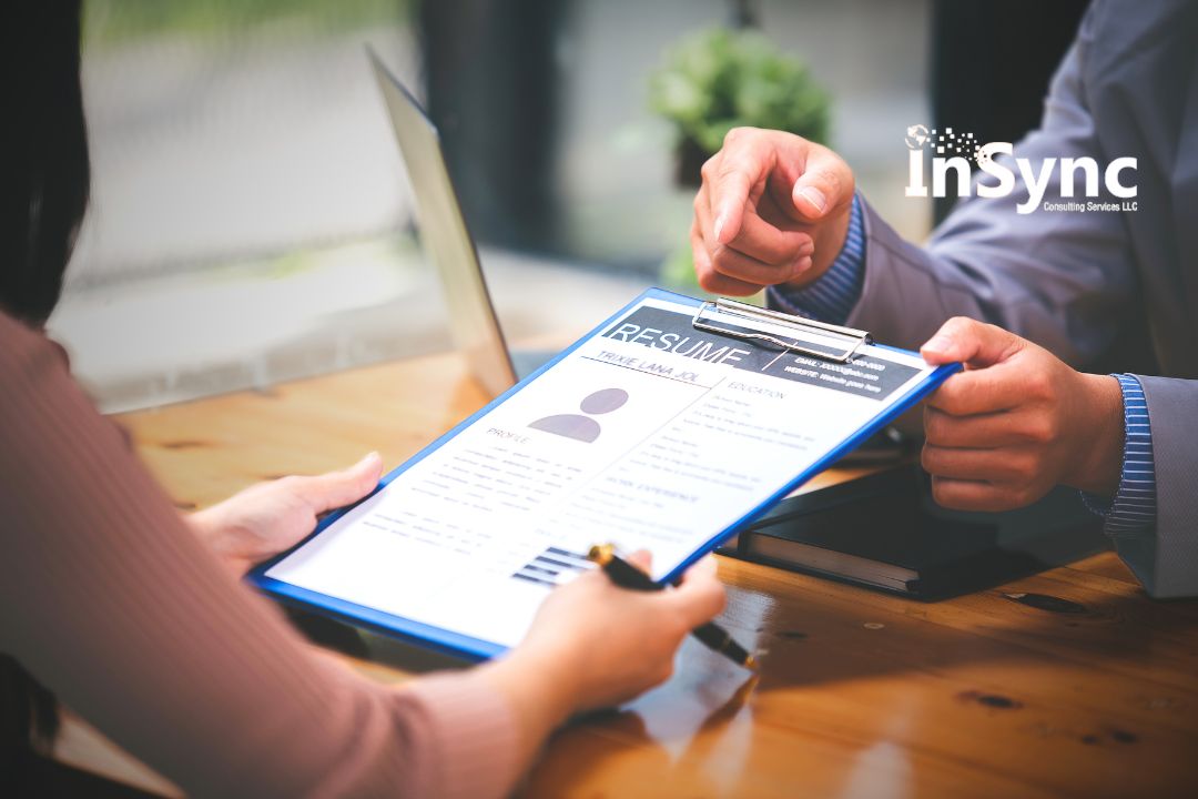 Two professionals reviewing a printed resume on a clipboard during a job interview at a wooden desk, with a laptop and the InSync Consulting Services logo visible in the background. Two professionals reviewing a printed resume on a clipboard during a job interview at a wooden desk, with a laptop and the InSync Consulting Services logo visible in the background.