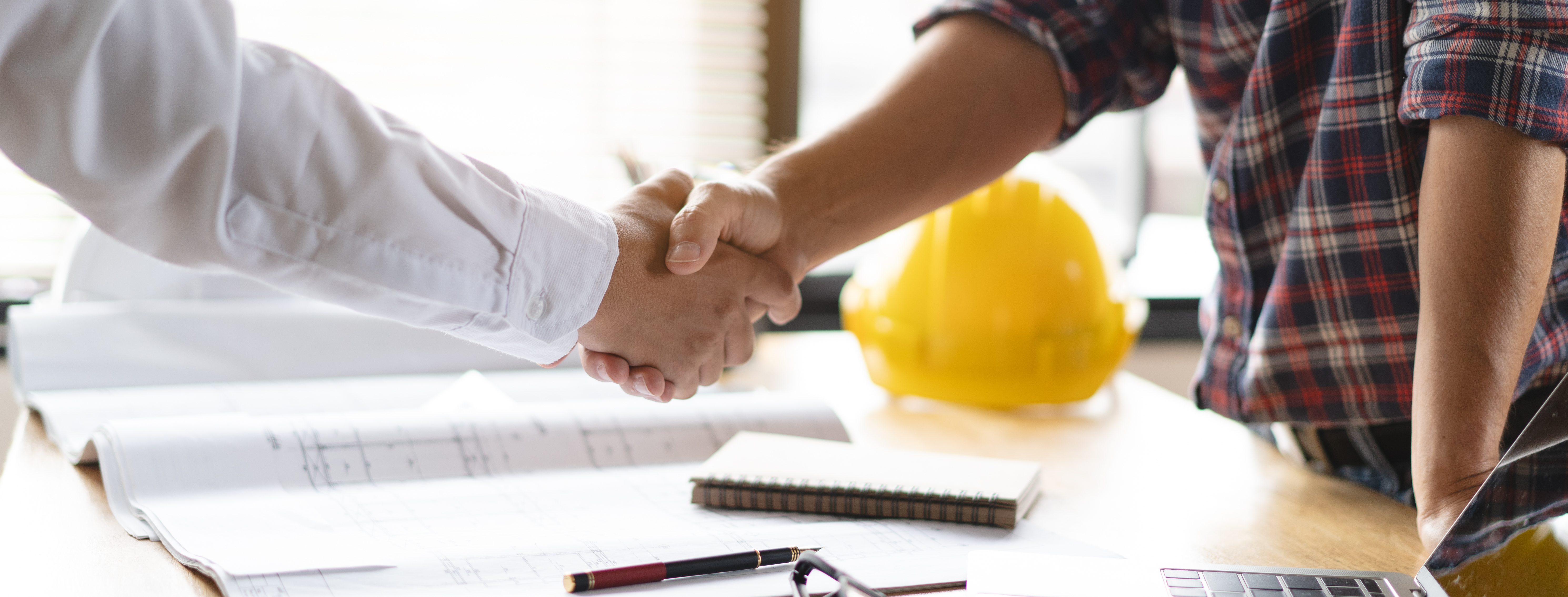 Contractor and homeowner shaking hands over construction plans with a hard hat on the table, symbolizing project approval and agreement.