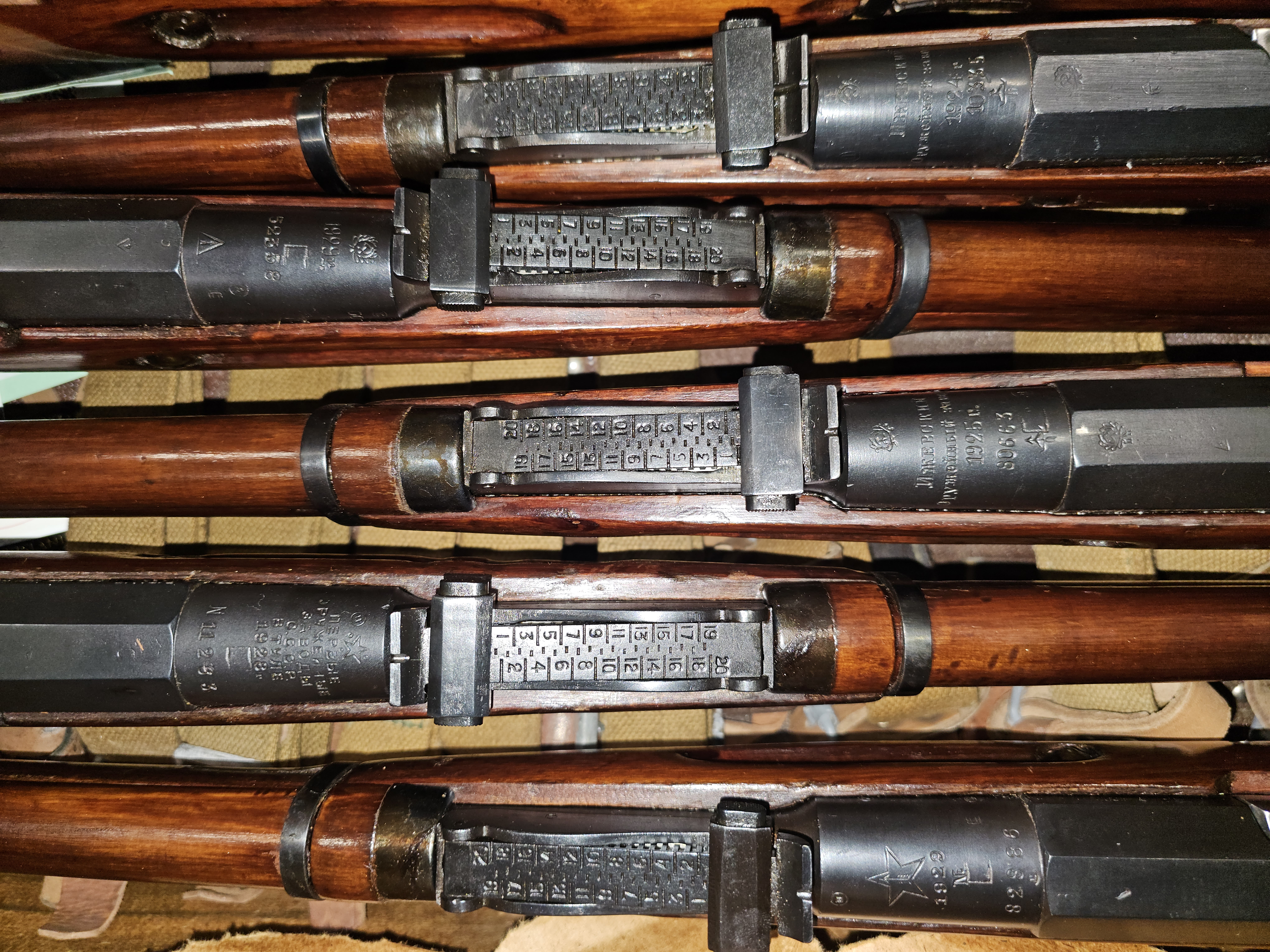 Close-up image showcasing the receiver markings, arsenal stamps (Izhevsk and Tula), serial numbers, and rear sights of multiple historic Soviet M91/30 Mosin-Nagant rifles arranged side by side. Close-up image showcasing the receiver markings, arsenal stamps (Izhevsk and Tula), serial numbers, and rear sights of multiple historic Soviet M91/30 Mosin-Nagant rifles arranged side by side.