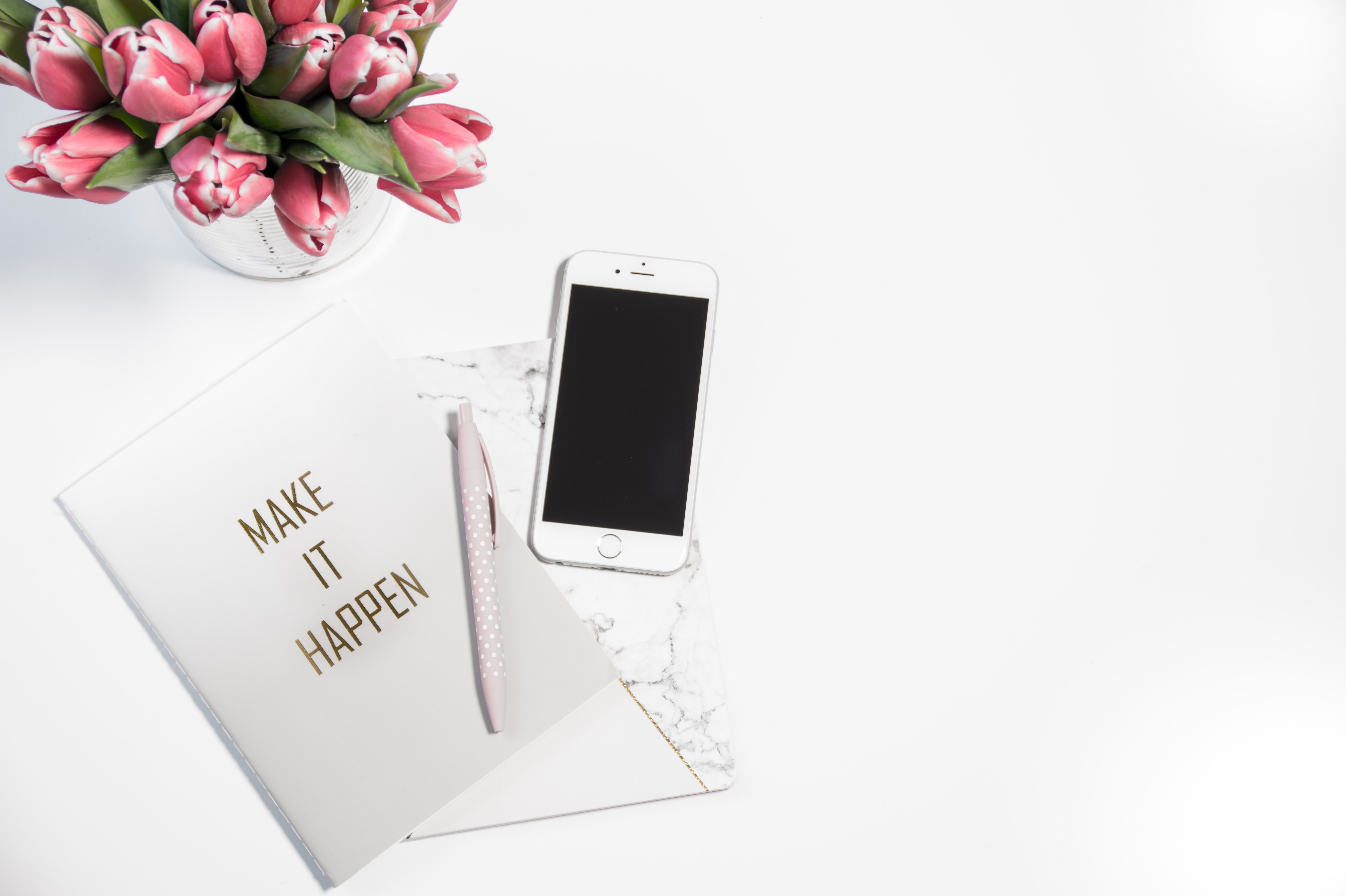 Image of a notebook with the text 'Make it happen' on a white desk with some pink flowers in a case