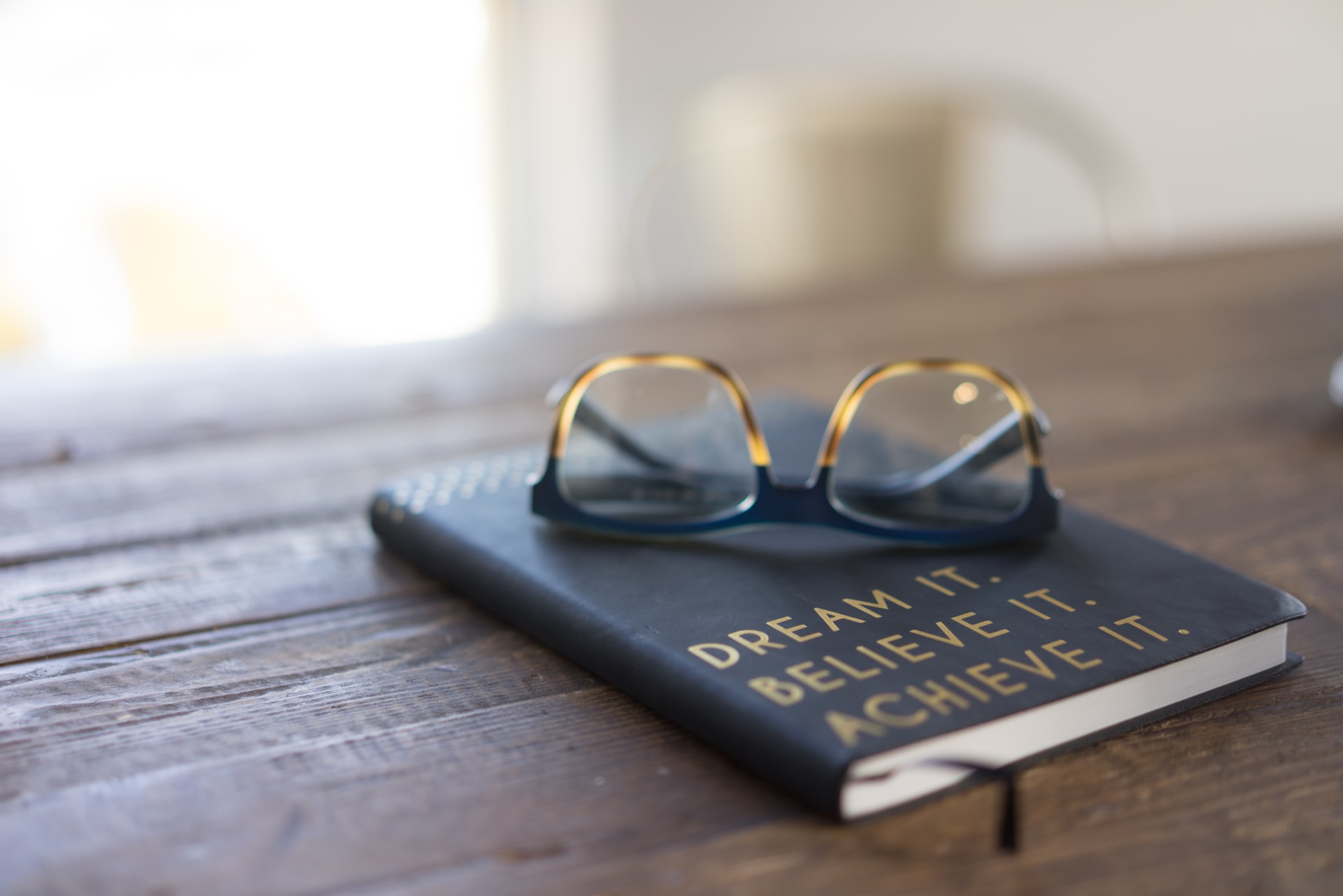 A book on a desk with a pair of the glasses on top. The book says 'Dream it. Believe it. Achieve it.