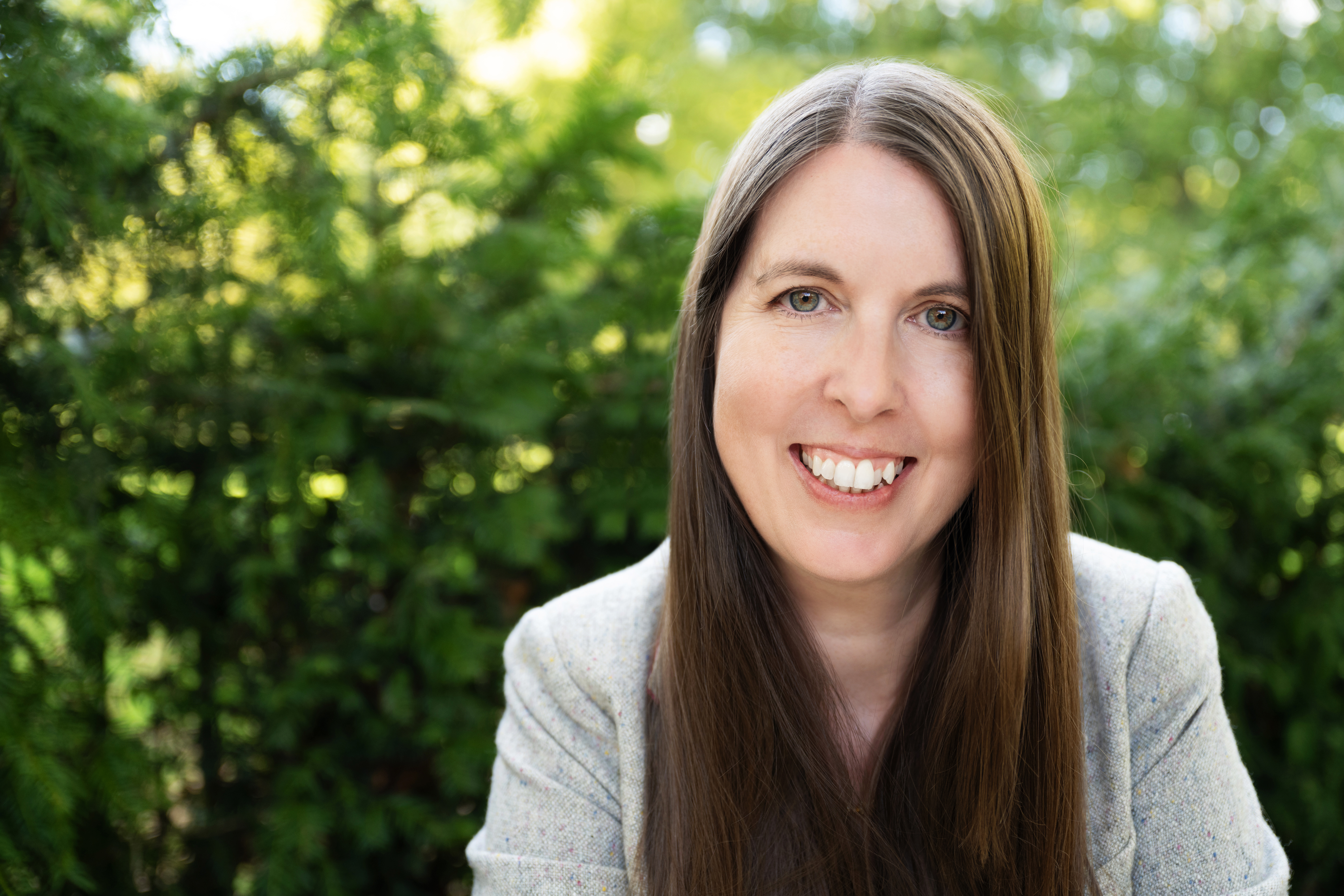 Photo of Sarah in a grey blazer smiling at the camera with green trees in the background