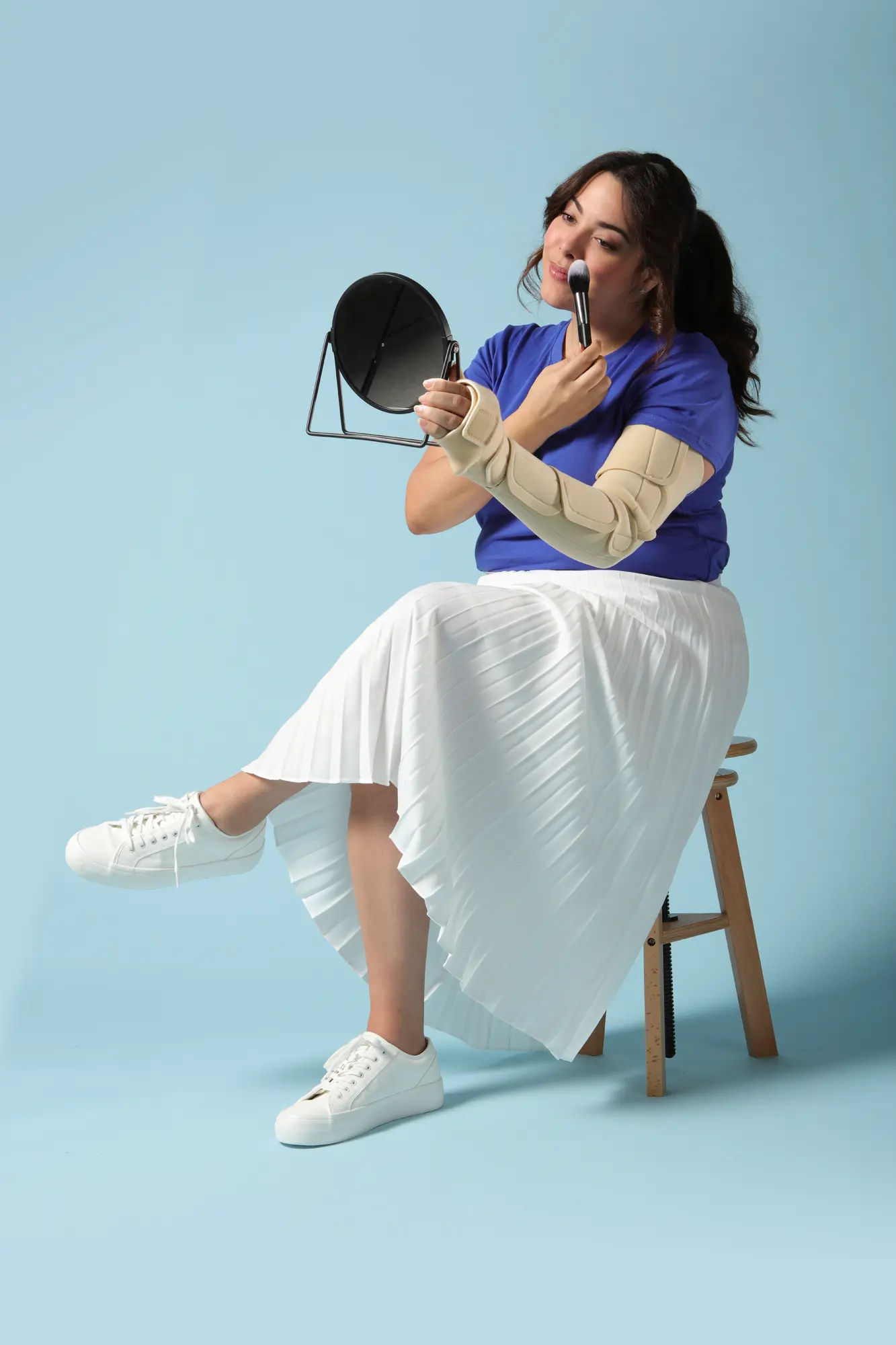 Woman demonstrating a beige adjustable compression arm wrap designed to support lymphatic health and everyday comfort, while applying makeup at home.