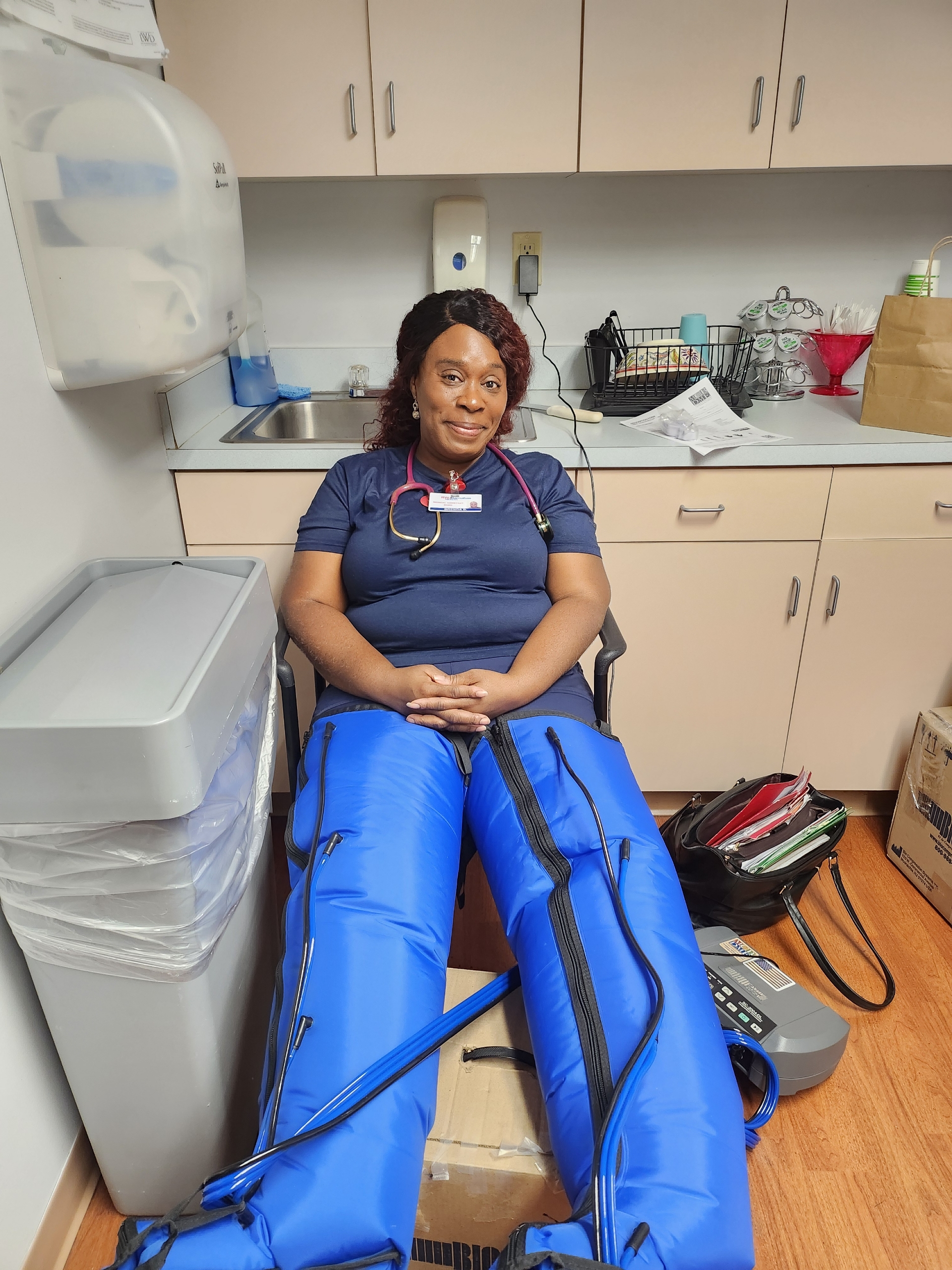 Nurse demonstrating the use of a pneumatic compression therapy device for leg circulation, wearing blue compression sleeves connected to a control unit.