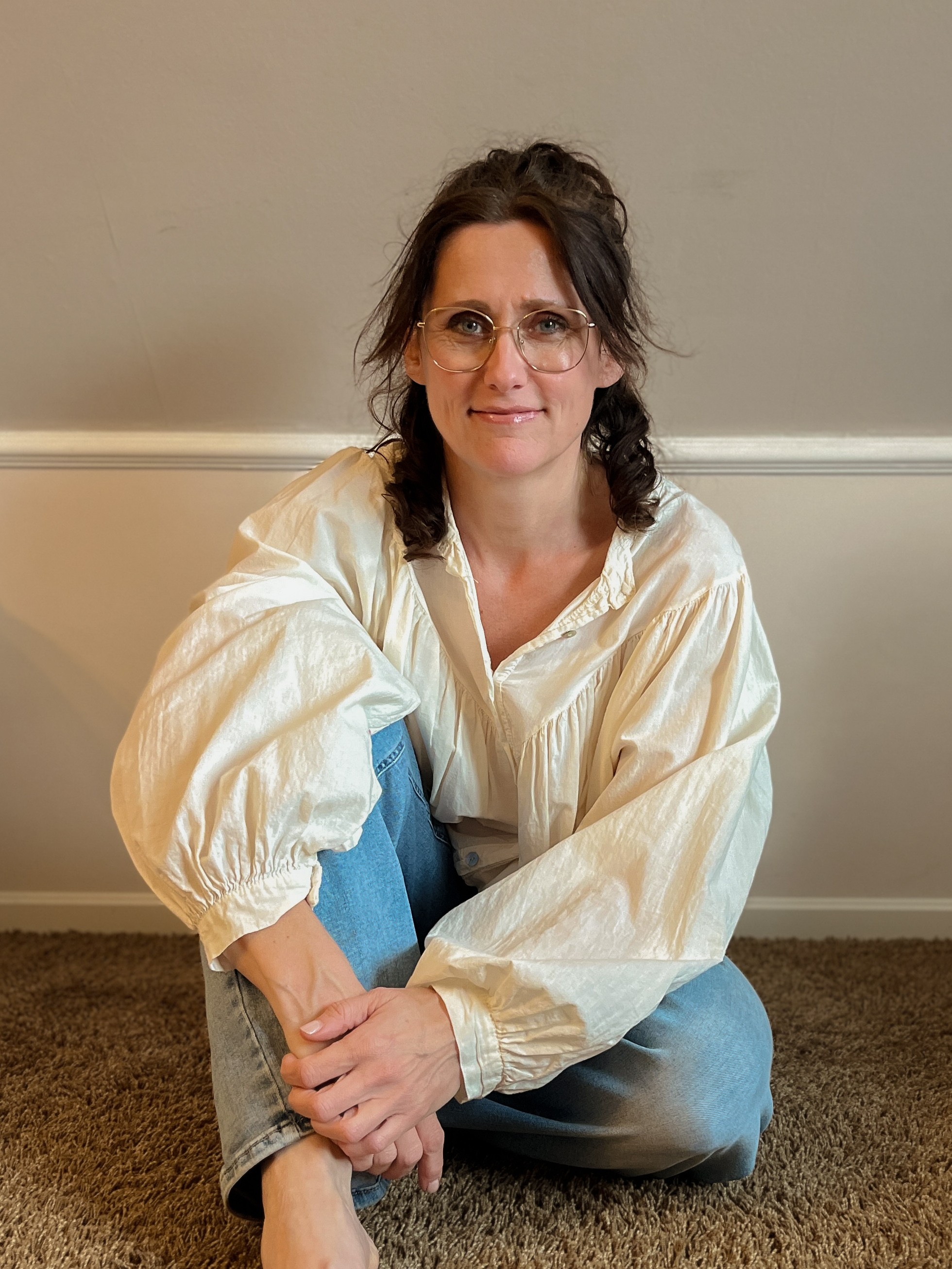 Foto Anja Glashörster, auf dem Boden sitzend, cremefarbene Bluse, weite blaue Jeans