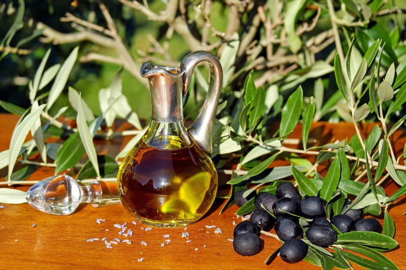 a glass bottle containing olive oil on a table with fresh olives and branches of olive