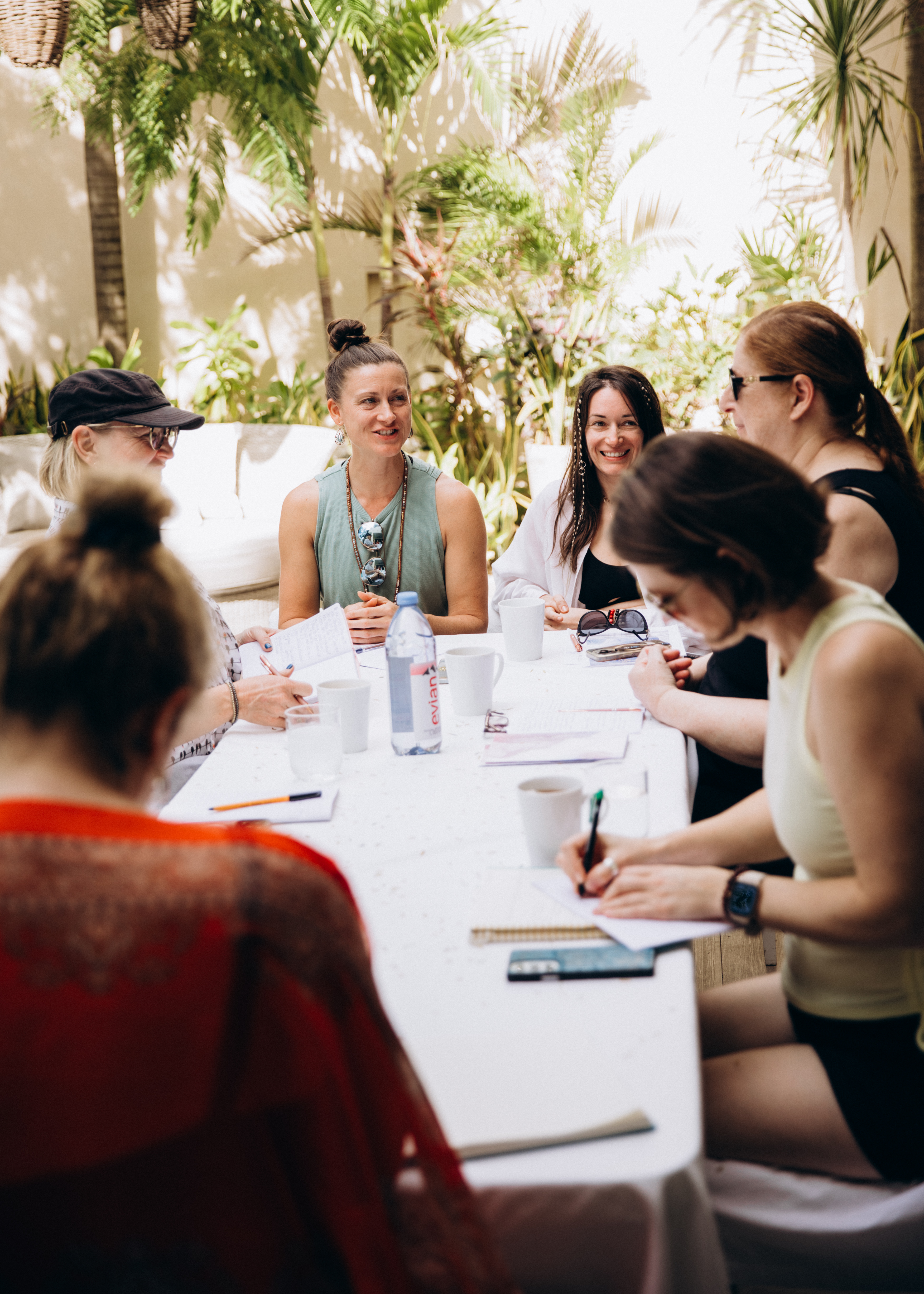 A group of women sitting outdoors at a white table during a retreat session, writing, sharing, and smiling while surrounded by greenery and natural light. A group of women sitting outdoors at a white table during a retreat session, writing, sharing, and smiling while surrounded by greenery and natural light.