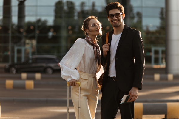 Charming man in sunglasses, black suit and attractive blonde woman in white blouse, beige pants.