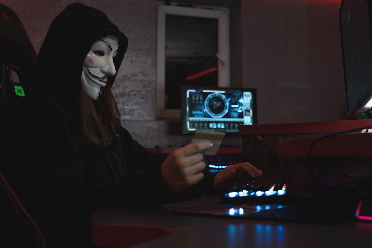 Masked man in black hoodie holding a white ID card with computer in background 