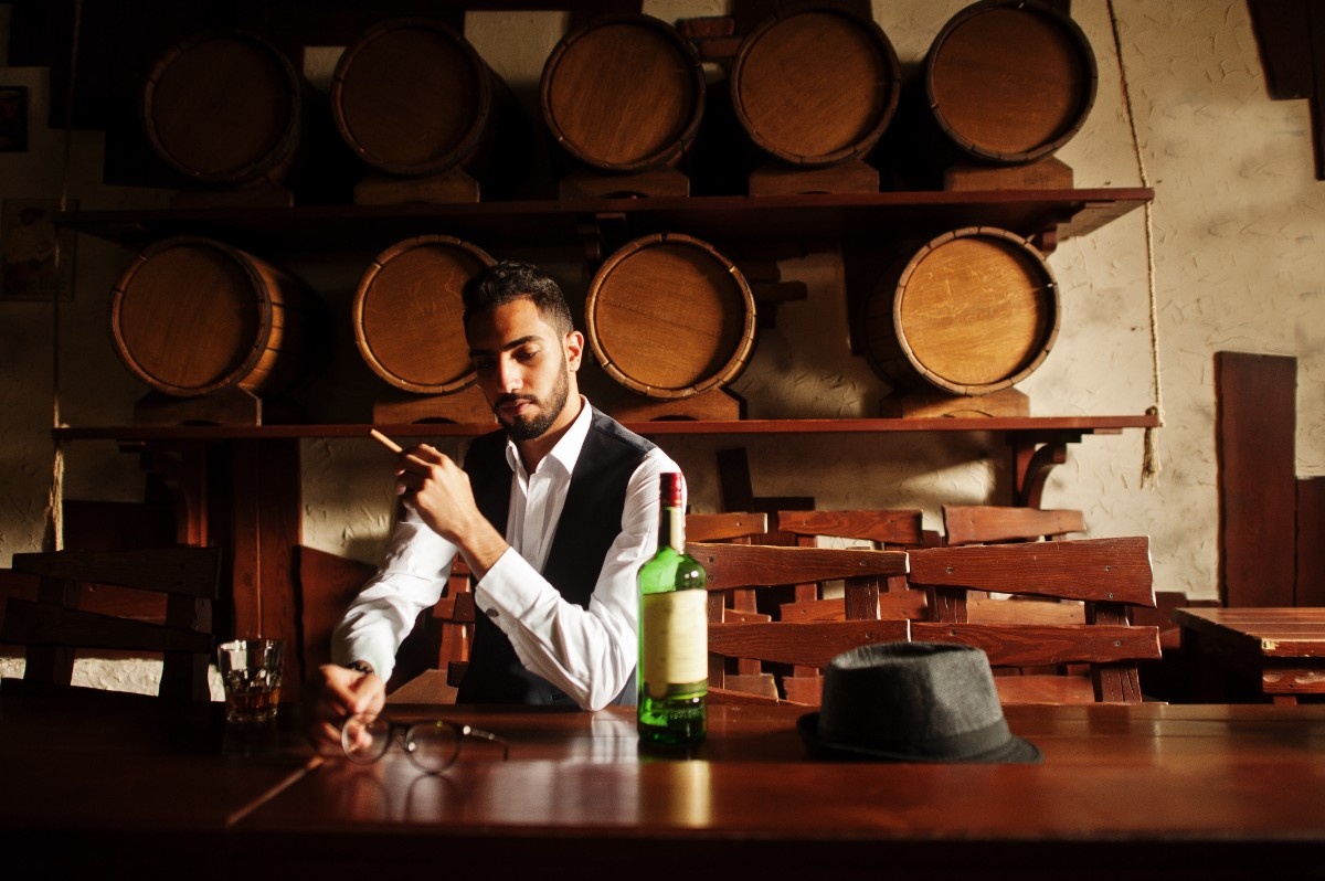 Man formally dressed holding cigar sitting down with wooden barrels behind him.