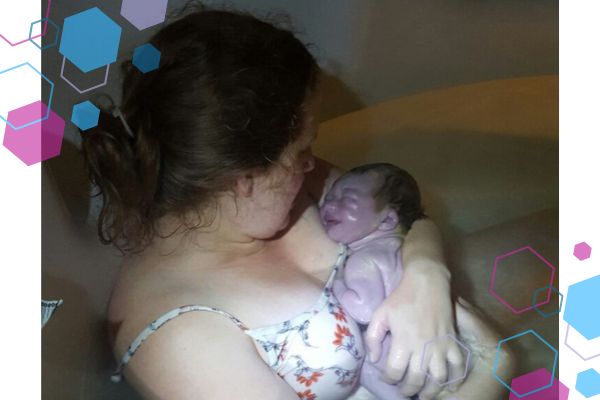 Jilly's positive birth after trauma. She is sitting in a birth pool cuddling her brand new baby daughter. Both mum and baby are looking at each other. The room is dark and the baby is so new she looks grey or purple.