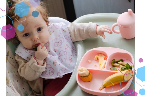 A baby sitting in a high chair looking at the camera, they have food in their hand and in their mouth and a plate of food next to them on the highchair tray A baby sitting in a high chair looking at the camera, they have food in their hand and in their mouth and a plate of food next to them on the highchair tray