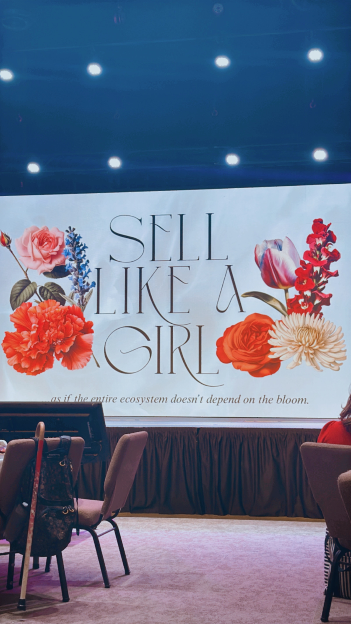 Stage backdrop at a conference featuring the phrase 'Sell Like a Girl' surrounded by bold floral designs, emphasizing feminine empowerment in business and sales.