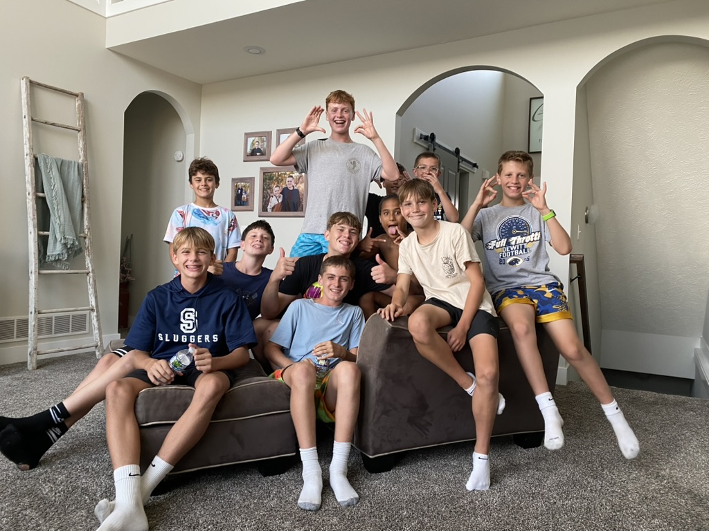 A group of boys playing in an outdoor pool with floats, a basketball hoop, and a slide, surrounded by fields and trees in the background. A group of boys playing in an outdoor pool with floats, a basketball hoop, and a slide, surrounded by fields and trees in the background.