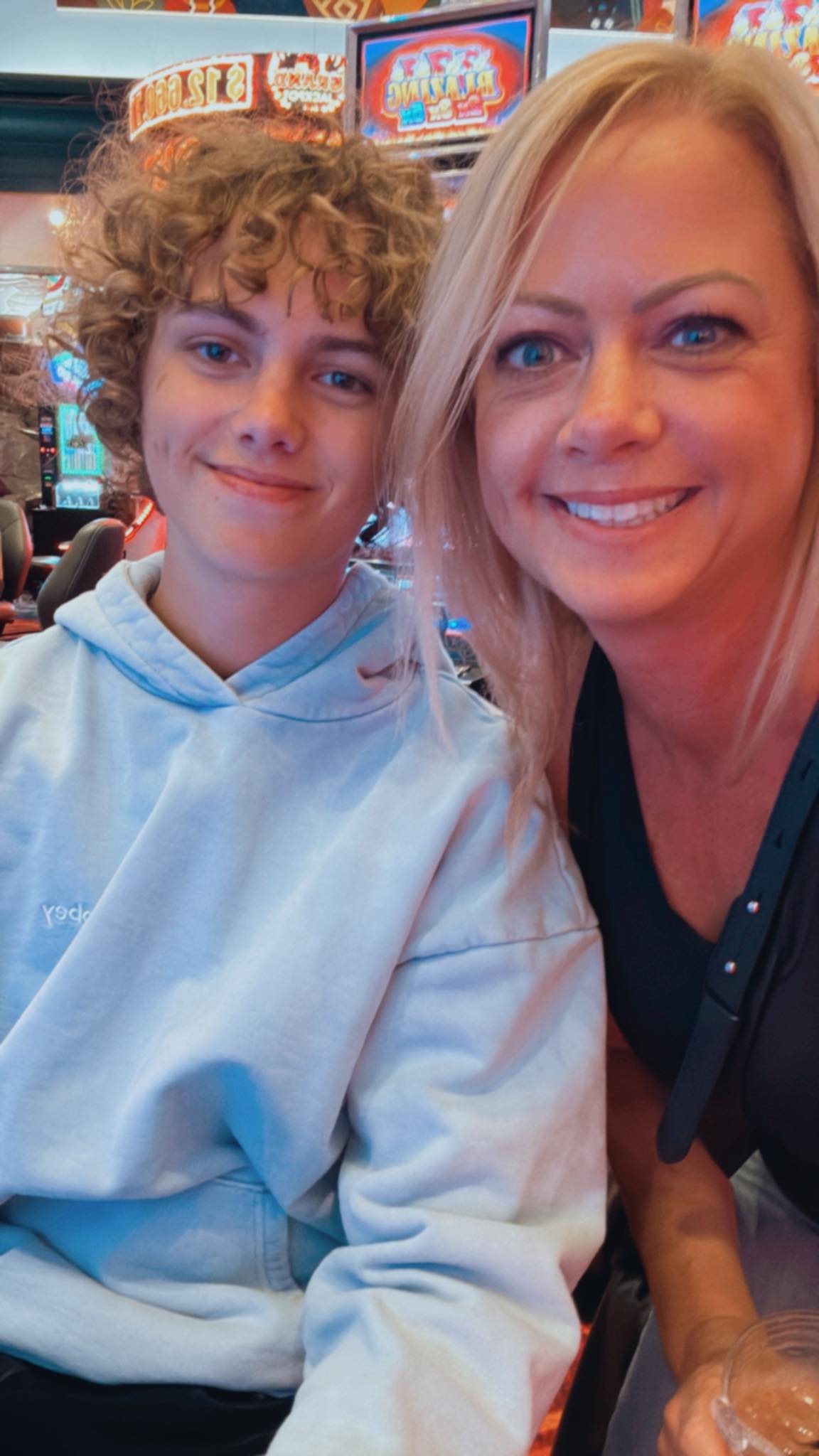 A smiling woman with straight blonde hair and blue eyes takes a selfie with a teenage boy who has curly light-brown hair and is wearing a pale blue hoodie. They’re sitting in what appears to be a casino or arcade with colorful slot machines and bright lights in the background. Both look happy and relaxed. A smiling woman with straight blonde hair and blue eyes takes a selfie with a teenage boy who has curly light-brown hair and is wearing a pale blue hoodie. They’re sitting in what appears to be a casino or arcade with colorful slot machines and bright lights in the background. Both look happy and relaxed.