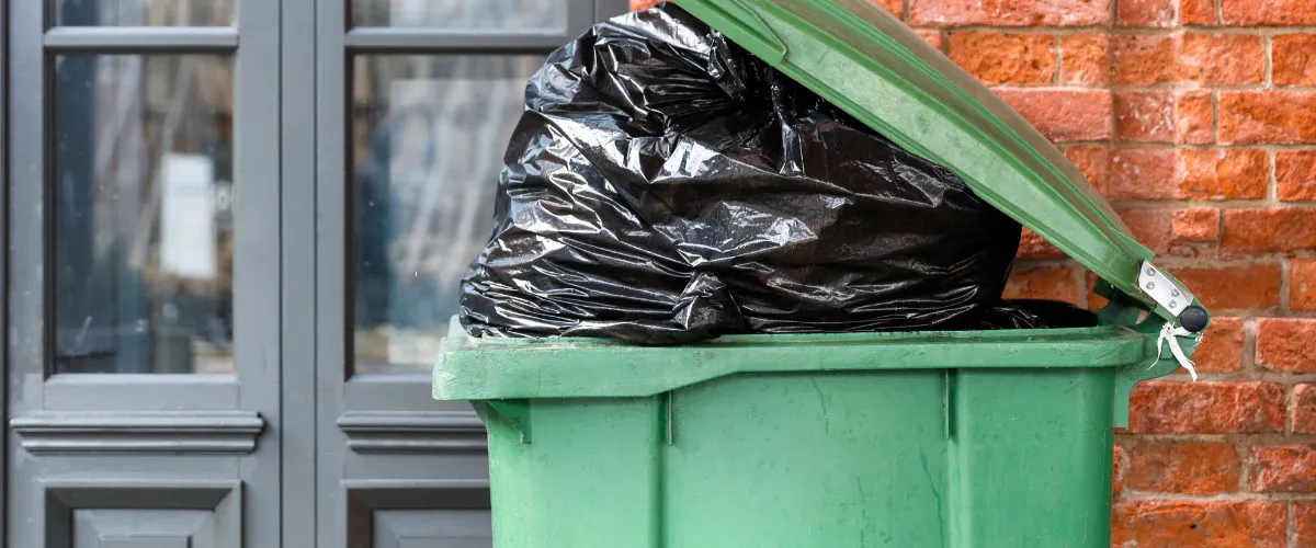 Green trash bin filled with black garbage bags placed outside a building with brick walls