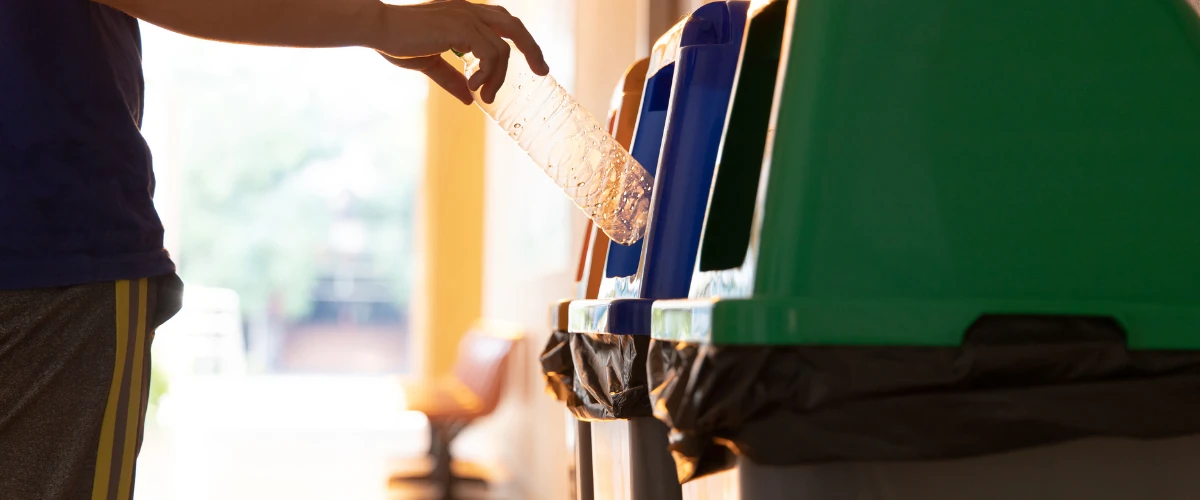 Person recycling a plastic bottle into a blue bin at a recycling station.
