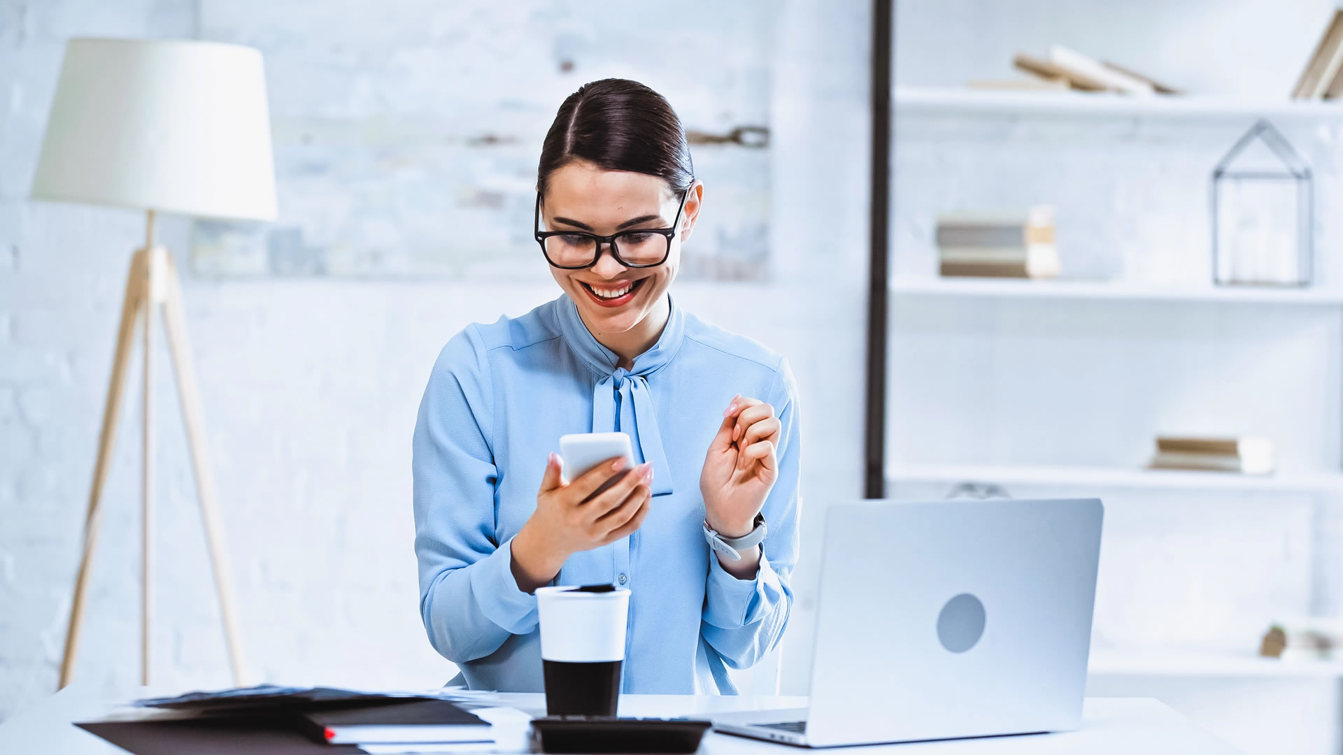 joyful-businesswoman-using-smartphone-coffee-laptop-workplace
