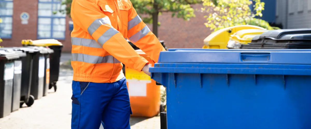 Waste management worker in orange uniform pushing a large blue commercial trash bin Waste management worker in orange uniform pushing a large blue commercial trash bin