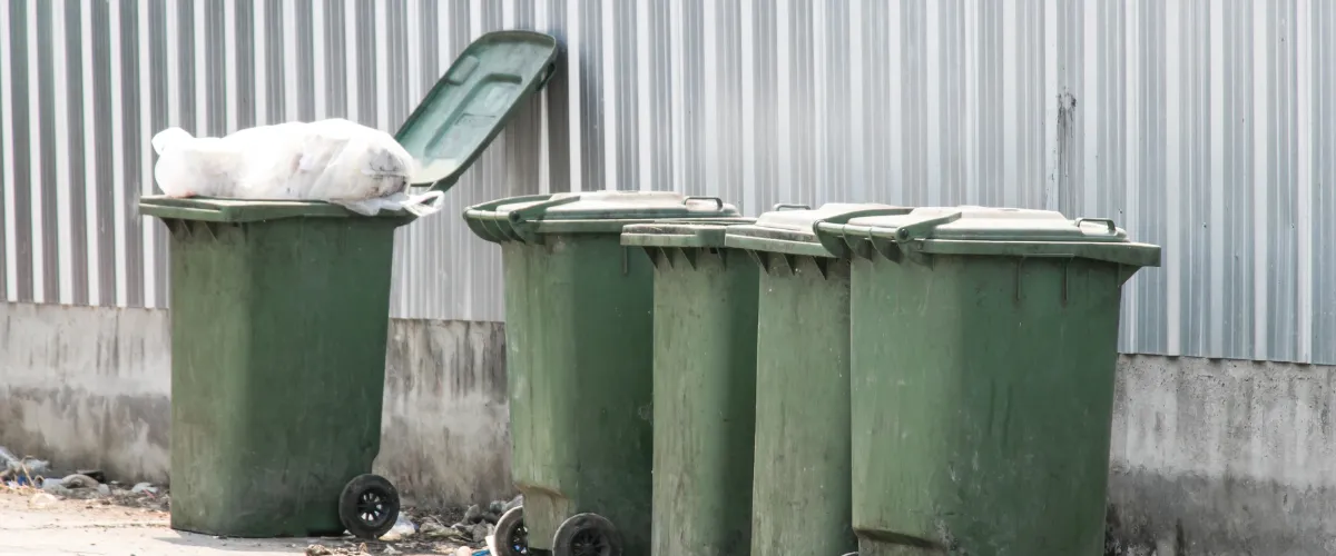 Outdoor green garbage bins lined up against a corrugated metal wall