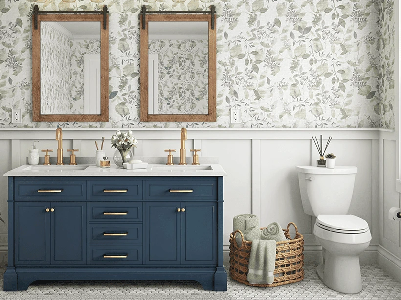 a bathroom with a blue vanity, double sinks, gold fixtures, two mirrors, floral wallpaper, a toilet, and a basket holding rolled towels and a plant.