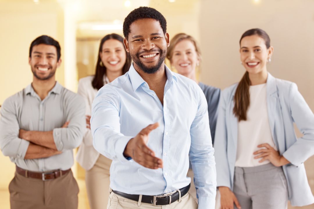 Confident businessman extending a handshake with a welcoming smile, standing in front of a diverse professional team, symbolizing trust and partnership in business. Confident businessman extending a handshake with a welcoming smile, standing in front of a diverse professional team, symbolizing trust and partnership in business.