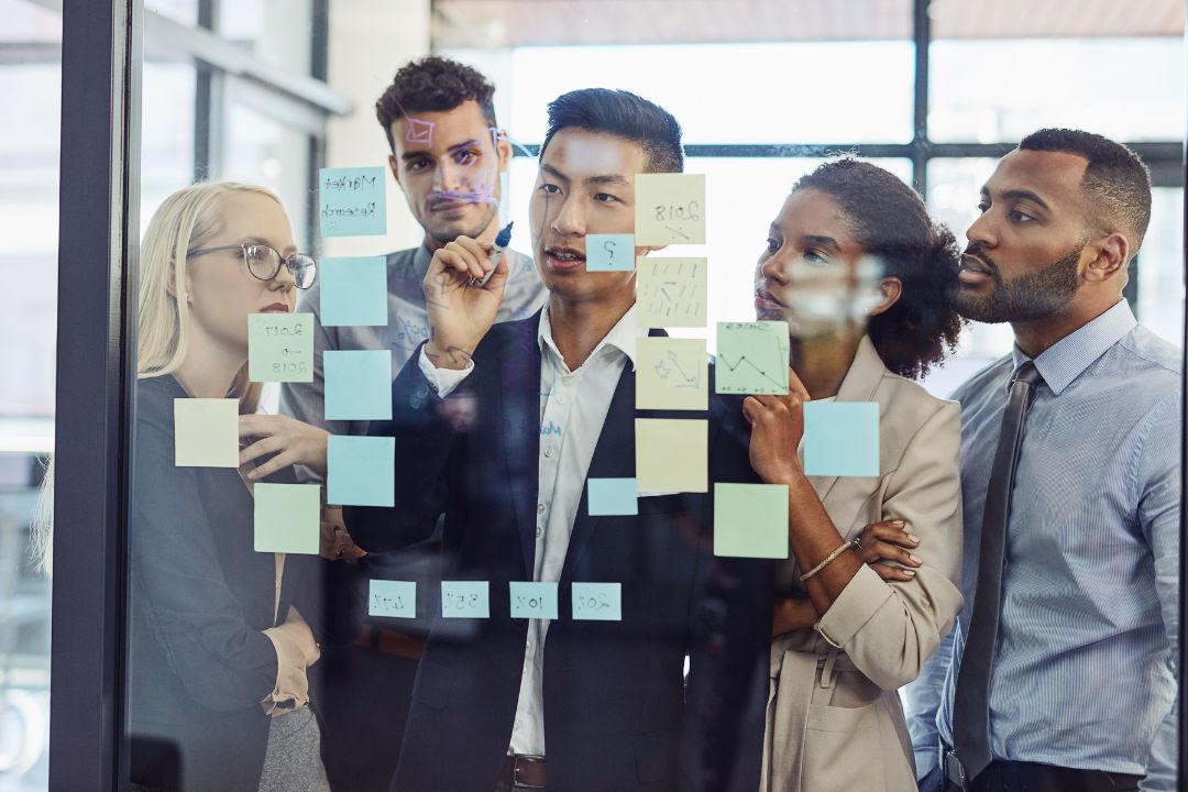 "Diverse team of business professionals brainstorming strategy on a glass board covered with sticky notes, discussing marketing plans and growth insights. "Diverse team of business professionals brainstorming strategy on a glass board covered with sticky notes, discussing marketing plans and growth insights.