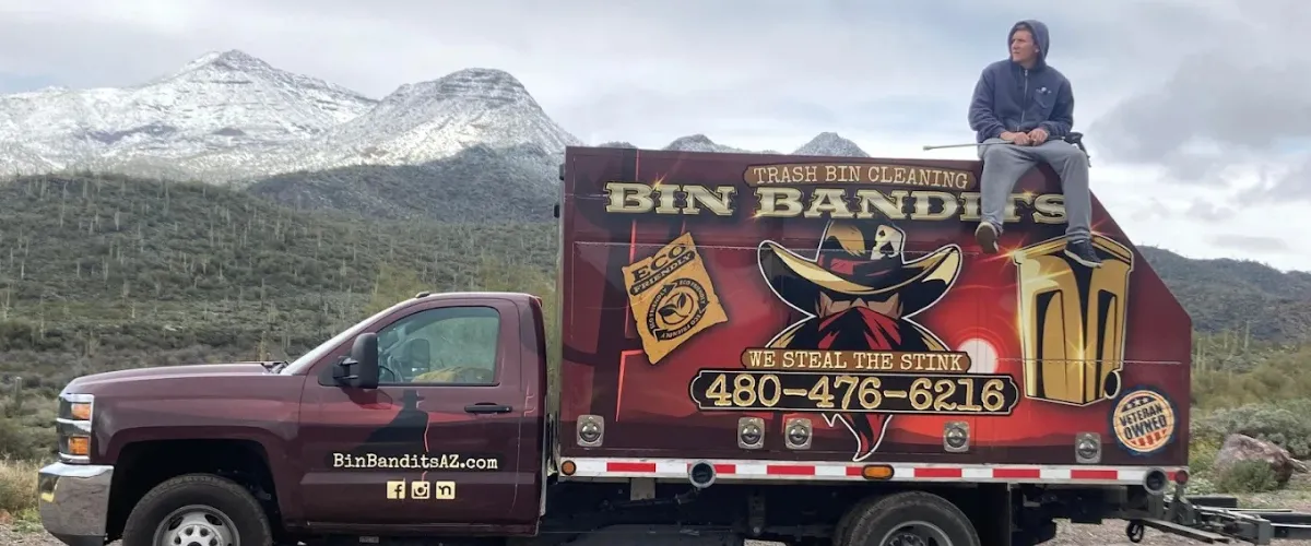 Side view of the Bin Bandits branded truck with a person sitting on top, set against a mountainous Arizona backdrop. Side view of the Bin Bandits branded truck with a person sitting on top, set against a mountainous Arizona backdrop.