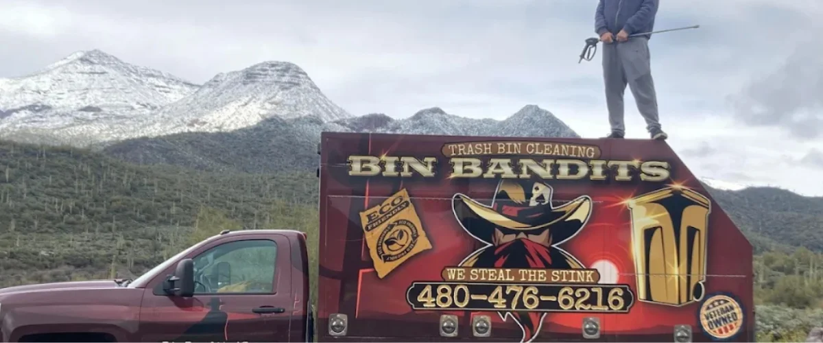 Bin Bandits trash bin cleaning truck with a worker standing on top, set against a mountain background Bin Bandits trash bin cleaning truck with a worker standing on top, set against a mountain background