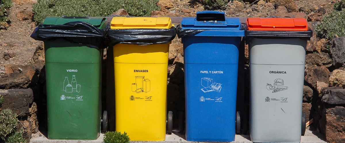 Four color-coded recycling bins for glass, plastic, paper, and organic waste lined up outdoors.