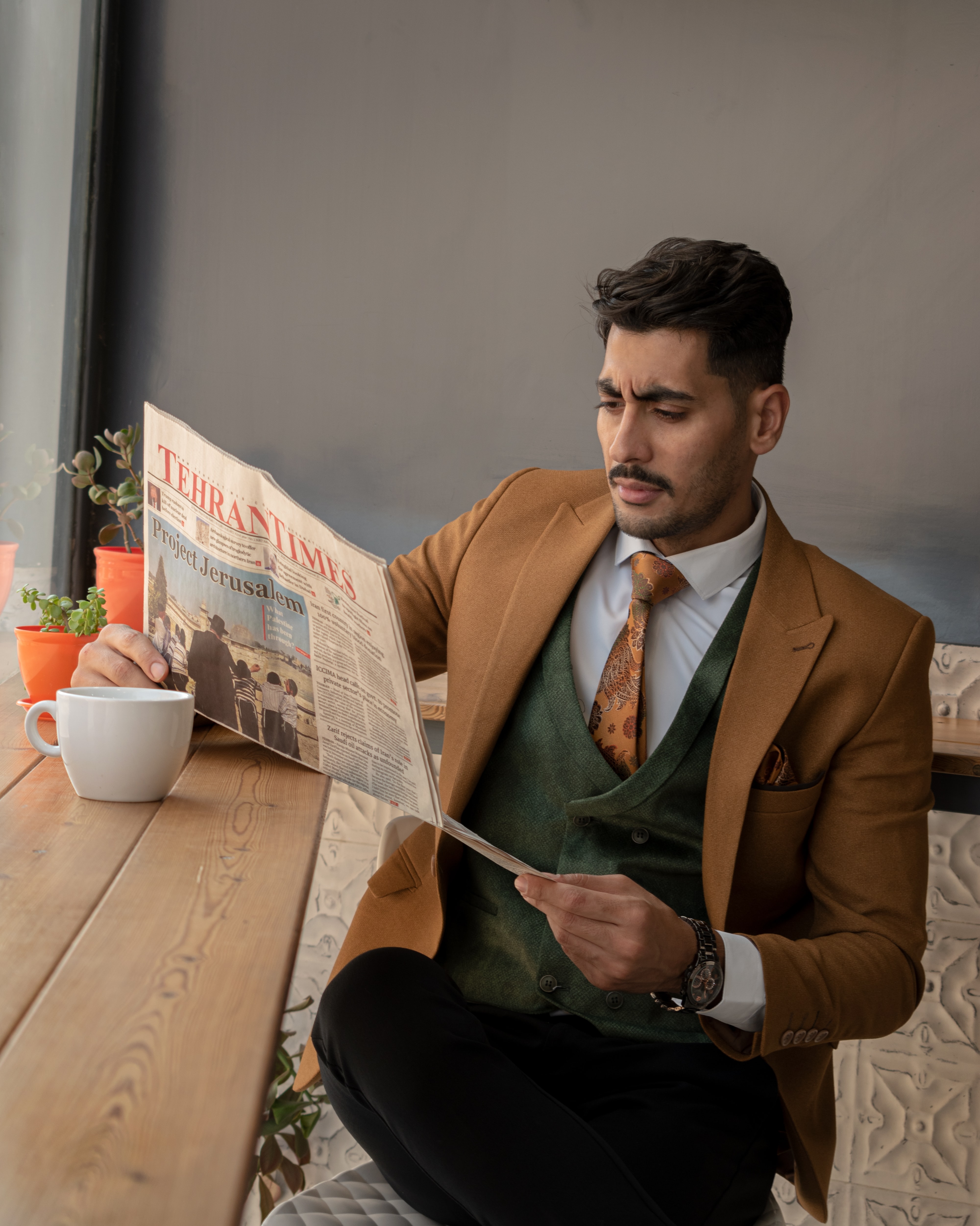 A man wearing a brown suit jacket, green vest and black dress pants reading a newspaper A man wearing a brown suit jacket, green vest and black dress pants reading a newspaper