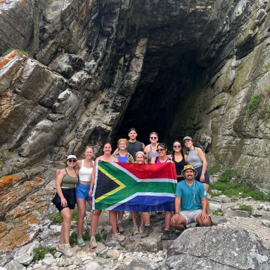 Group of travelers holding South African flag, standing in front of waterfall.