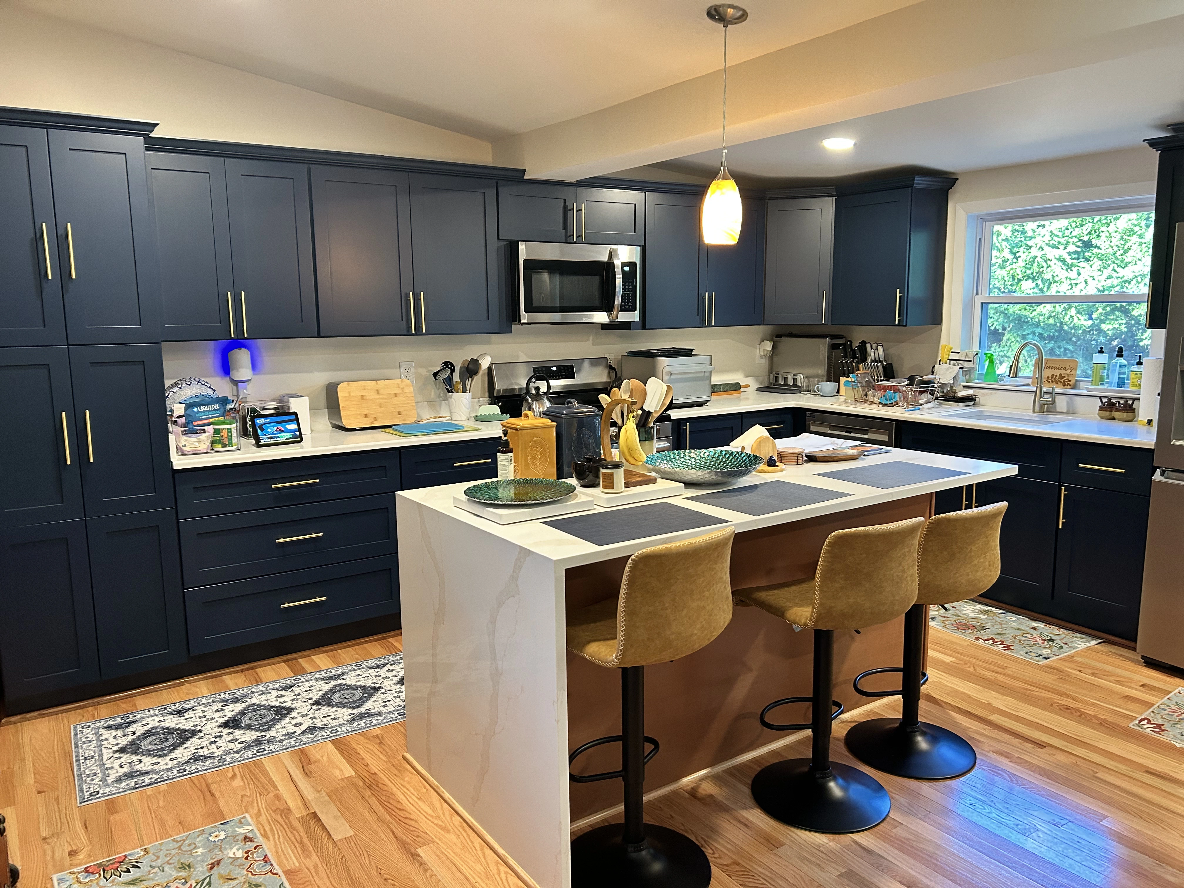 Modern kitchen with navy cabinets, white countertops, and bar stools, featuring various utensils and a sunny window view.