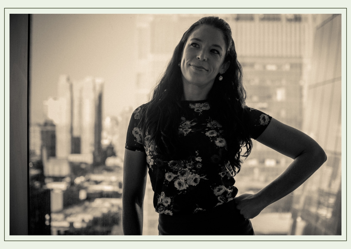 Black-and-white portrait of Monique, standing confidently in front of a large window with a city skyline blurred in the background. She has long dark hair, wears a floral top, and has a thoughtful, slightly playful expression, with one hand on her hip.
