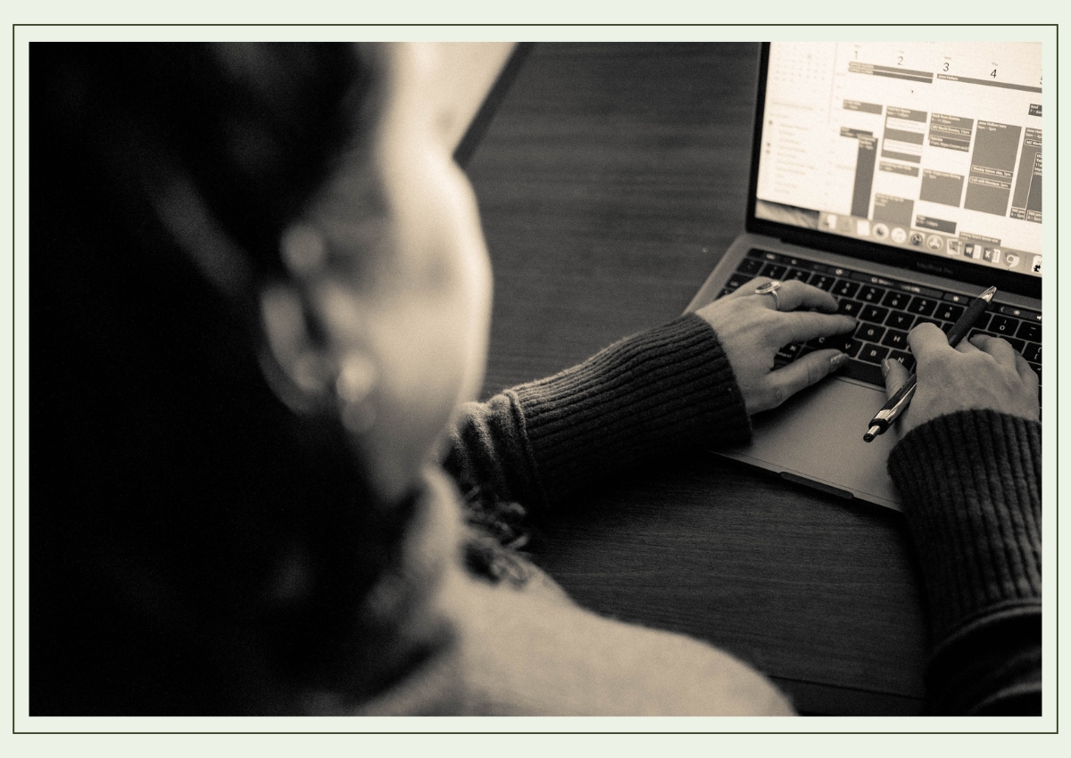 Black-and-white portrait of Monique, standing confidently in front of a large window with a city skyline blurred in the background. She has long dark hair, wears a floral top, and has a thoughtful, slightly playful expression, with one hand on her hip.