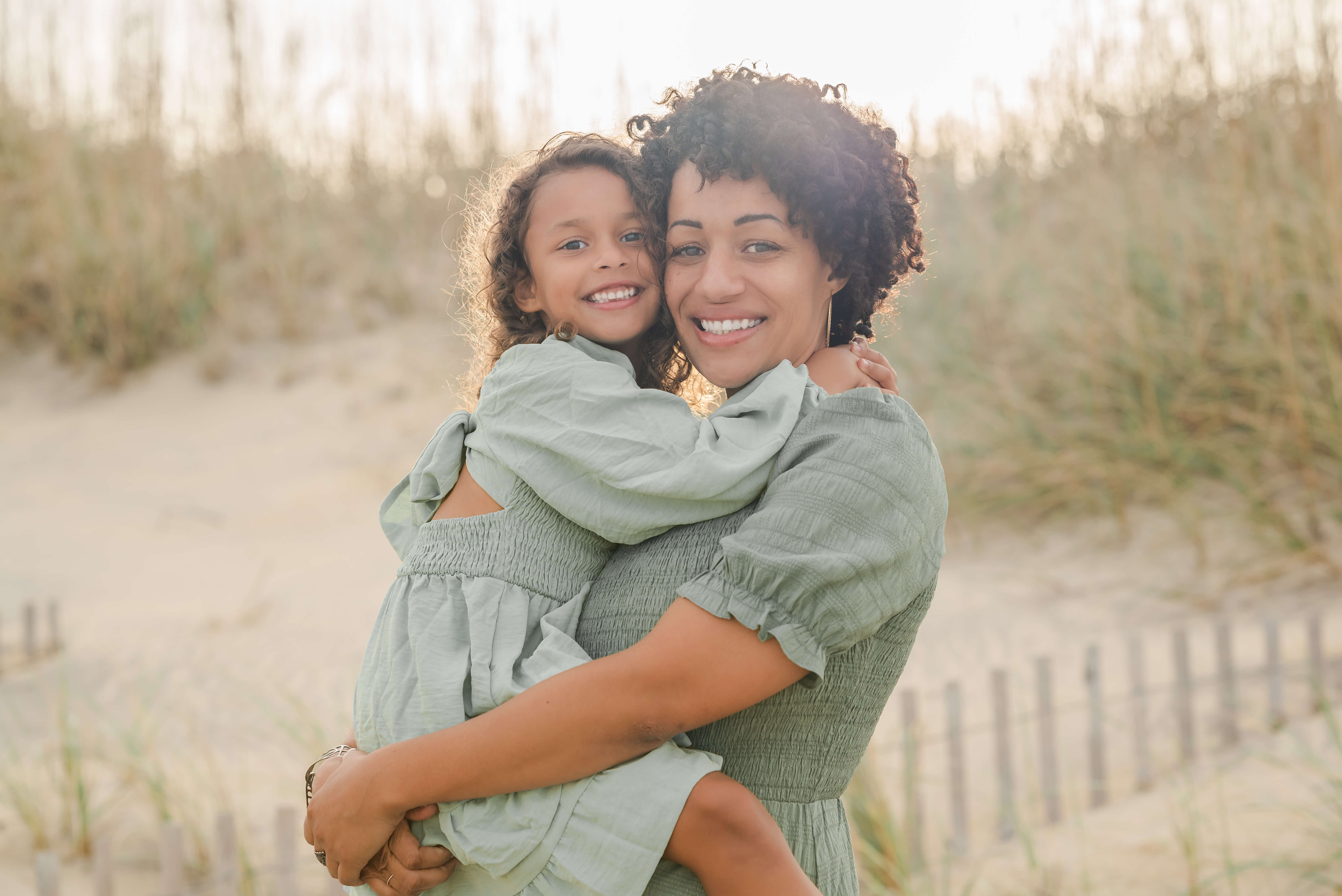 A mother holds her daughter in her arms. Both are wearing green dresses, which go well with the grassy dunes behind them on the beach. The sun is shining in their hair. A mother holds her daughter in her arms. Both are wearing green dresses, which go well with the grassy dunes behind them on the beach. The sun is shining in their hair.