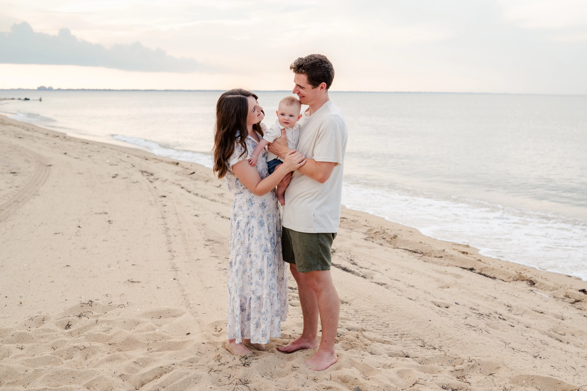 A mother and father look at each other while holding their baby boy between them. Their beach session was captured by Virginia Beach photographer Justine Renee Photography