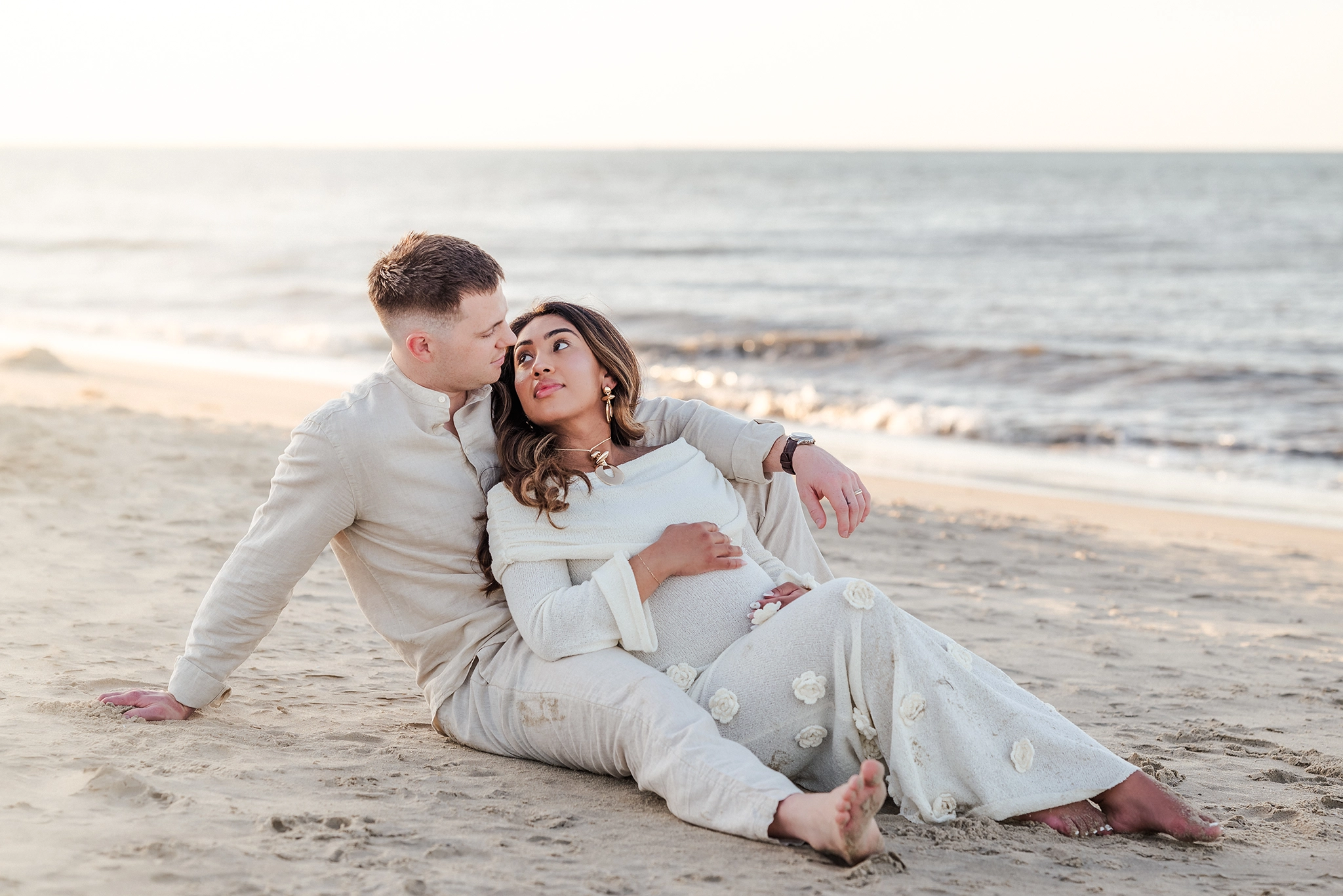 An expectant mother in a beige sweater dress, leans against her husband, who is also in neutral colors. They are at Fort Story beach in Virginia Beach for their session.
