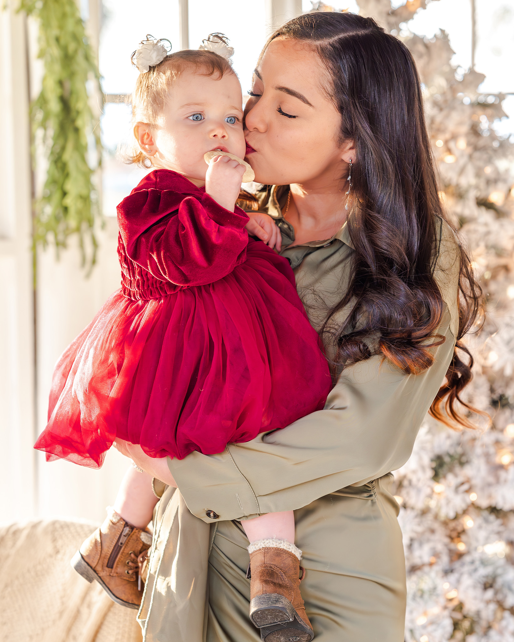 A mother in a green satin dress kisses her baby, who is in a red velvet dress, on the cheek during a Christmas mini session in Chesapeake, VA