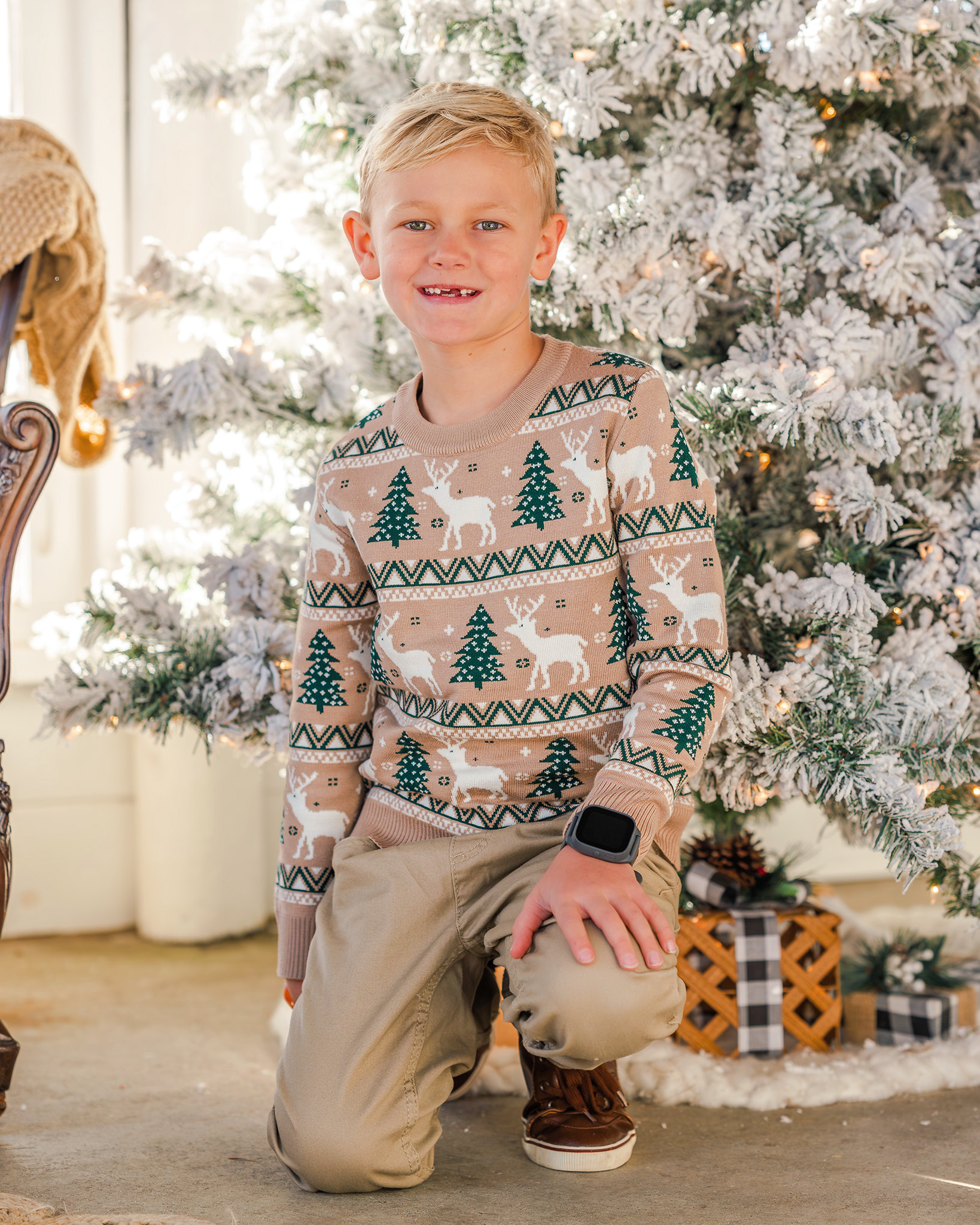 A child in a beige Christmas sweater sits in front of a frosted Christmas tree.